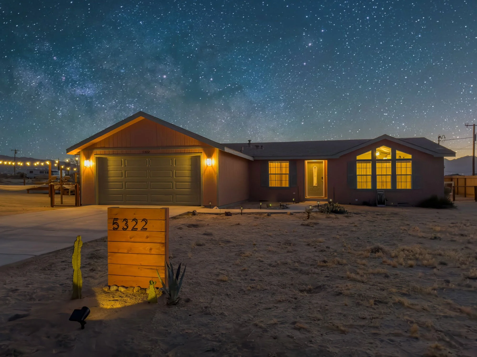 Night view of a house with a lit front door and large illuminated windows, a garage with lights on, and a yard with desert landscaping under a starry sky.