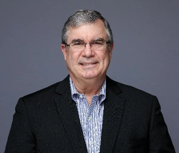 A middle-aged man with gray hair, glasses, and a smile, wearing a dark blazer and a patterned shirt, posing against a gray background.