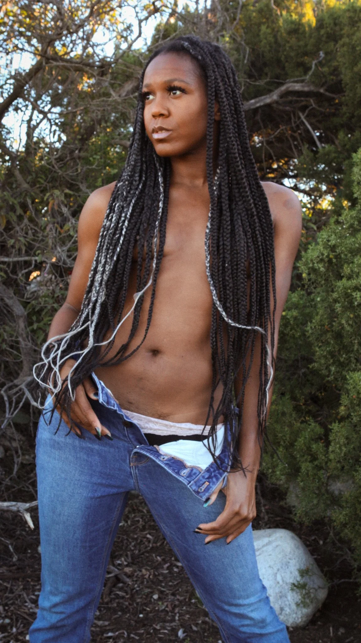 A woman with long braided hair standing outdoors among trees, wearing jeans and pulling them down slightly to reveal a black and white striped underwear.