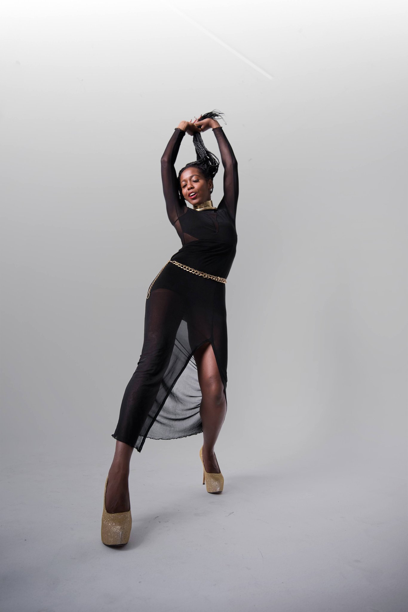 A woman dancing in a black sheer dress with a slit, gold heels, and gold jewelry, posing with arms raised above her head against a plain gray background.