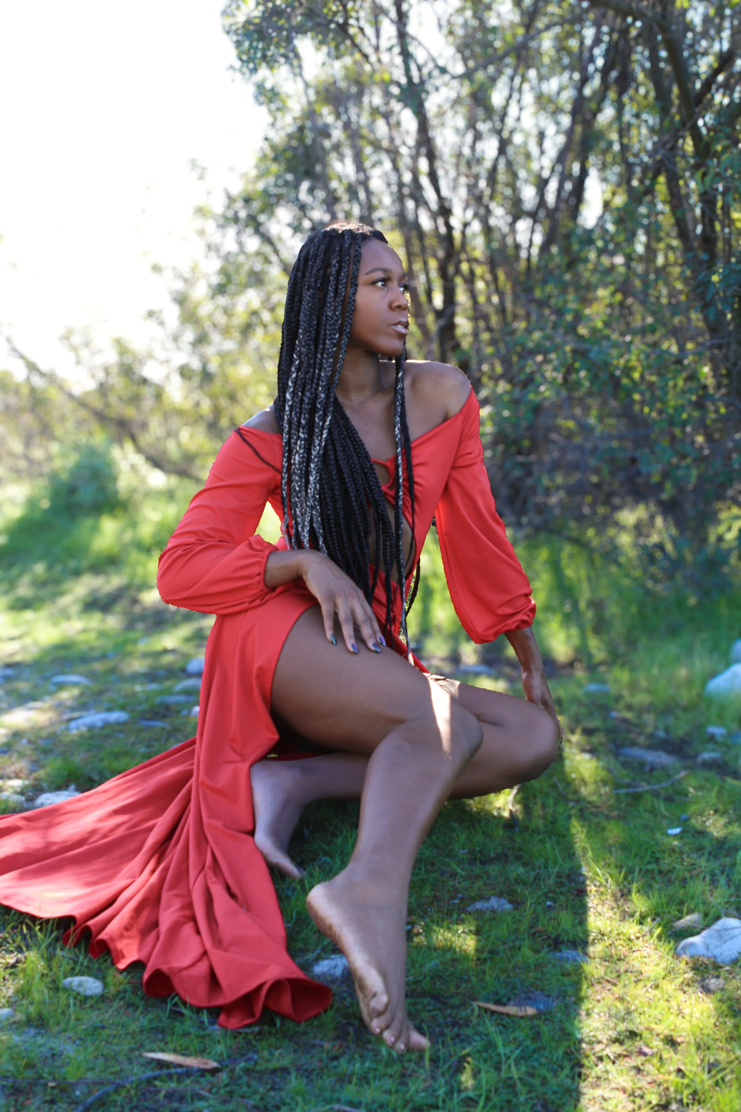 A woman with long braids wearing a red dress outdoors on grass, with a backdrop of trees and sunlight filtering through the leaves.