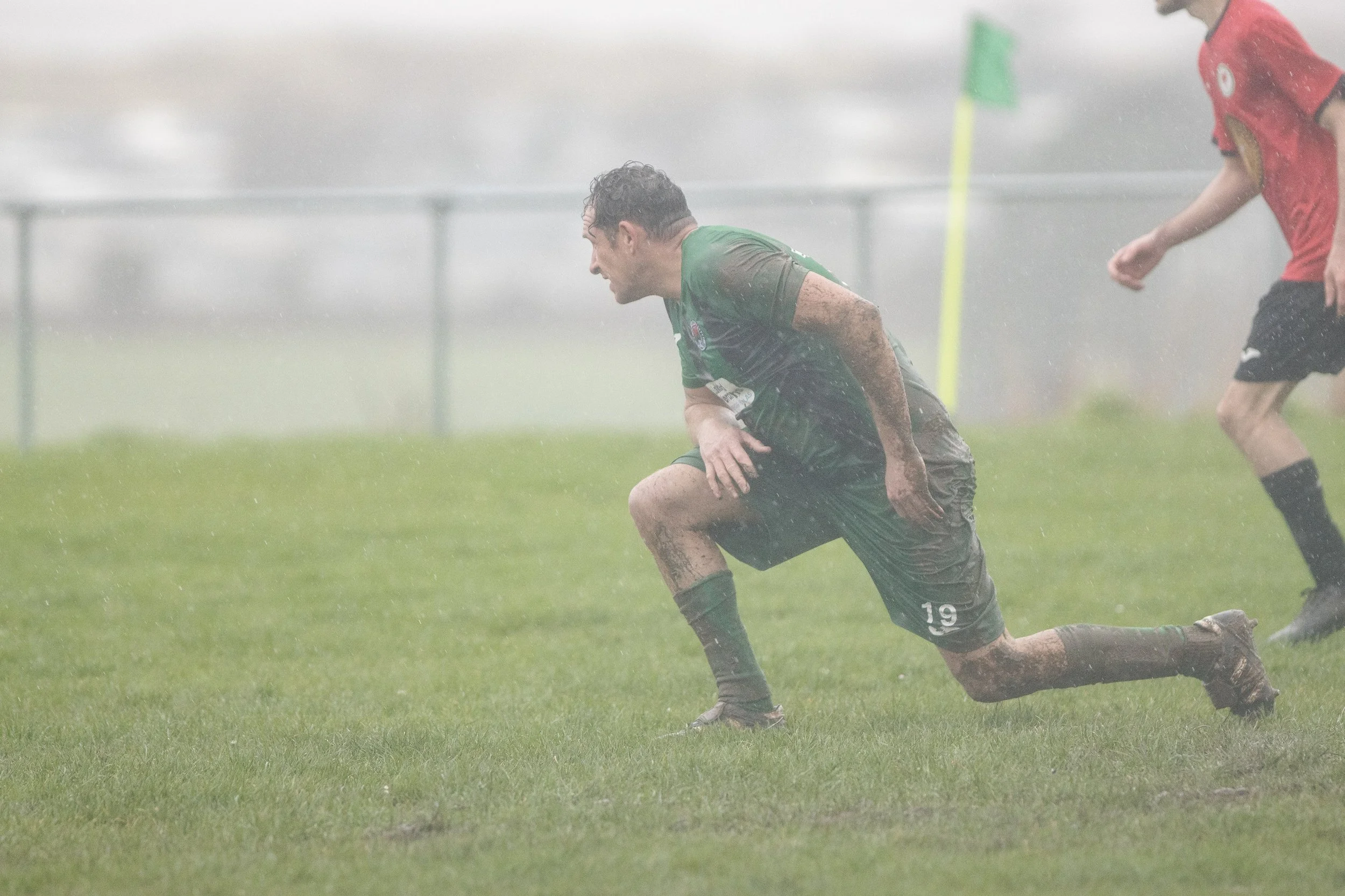 A soccer player in green and black uniform crawling on the muddy field during a game in rainy weather.