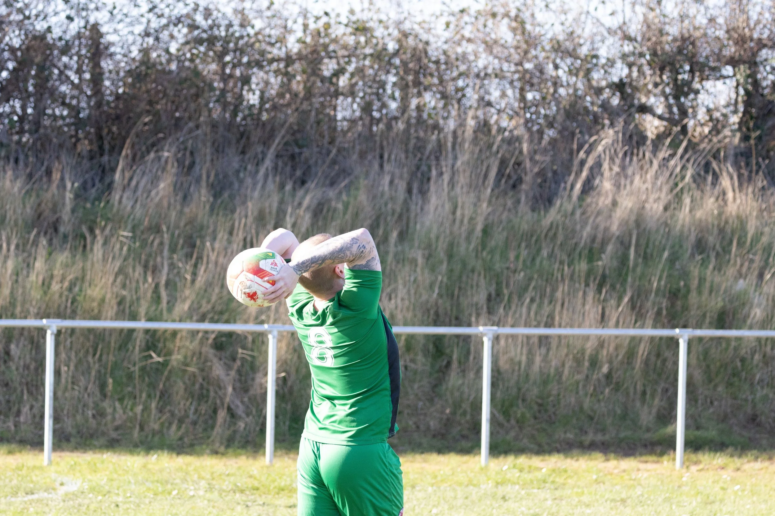 A soccer player in a green uniform is preparing to throw in the ball during a game, with a backdrop of dry grass and trees.