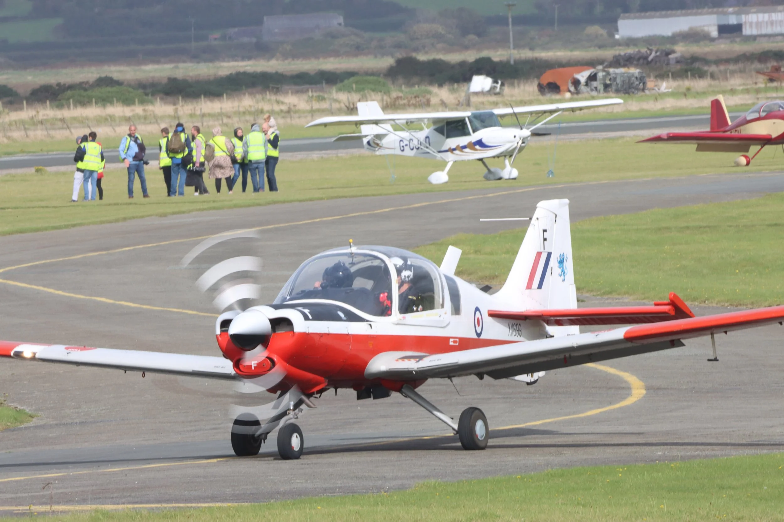 A red and white small propeller airplane on the runway with several groups of people in reflective safety vests and two small parked planes in the background.