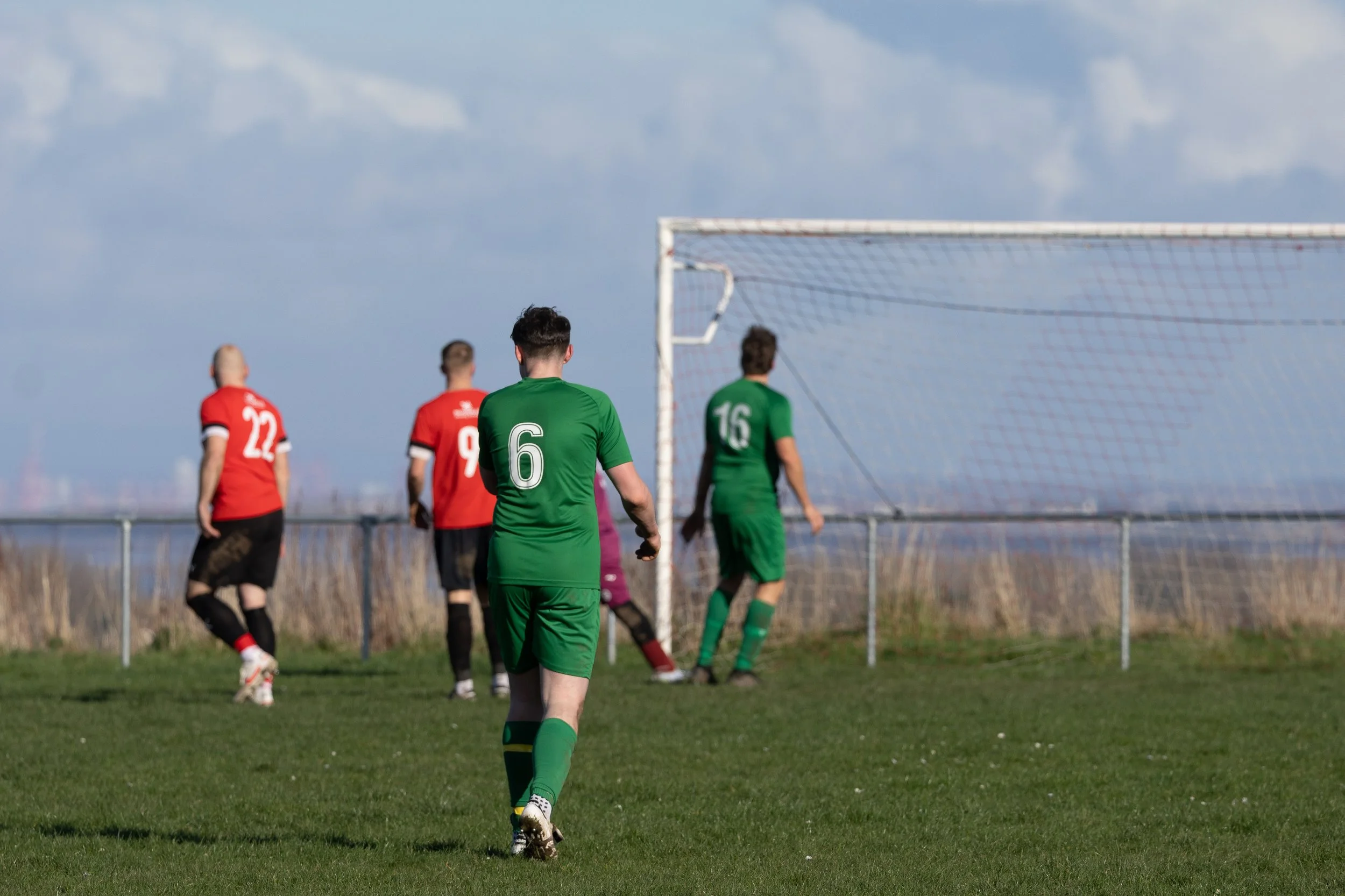Soccer players on a field, wearing red and green jerseys, near the goal during a match.