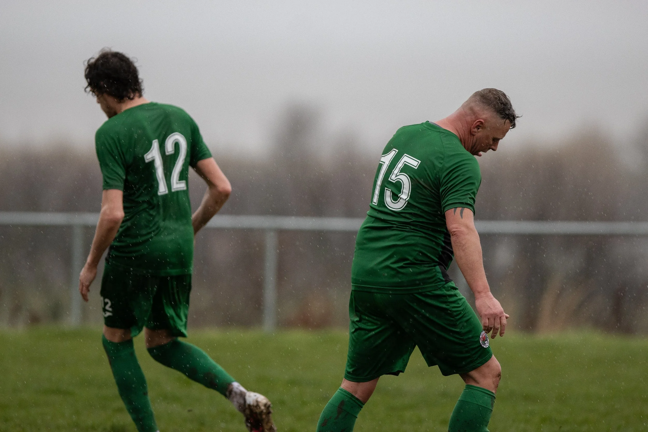Two soccer players in green jerseys standing on a rainy field, with one wearing number 12 and the other number 15, looking down.