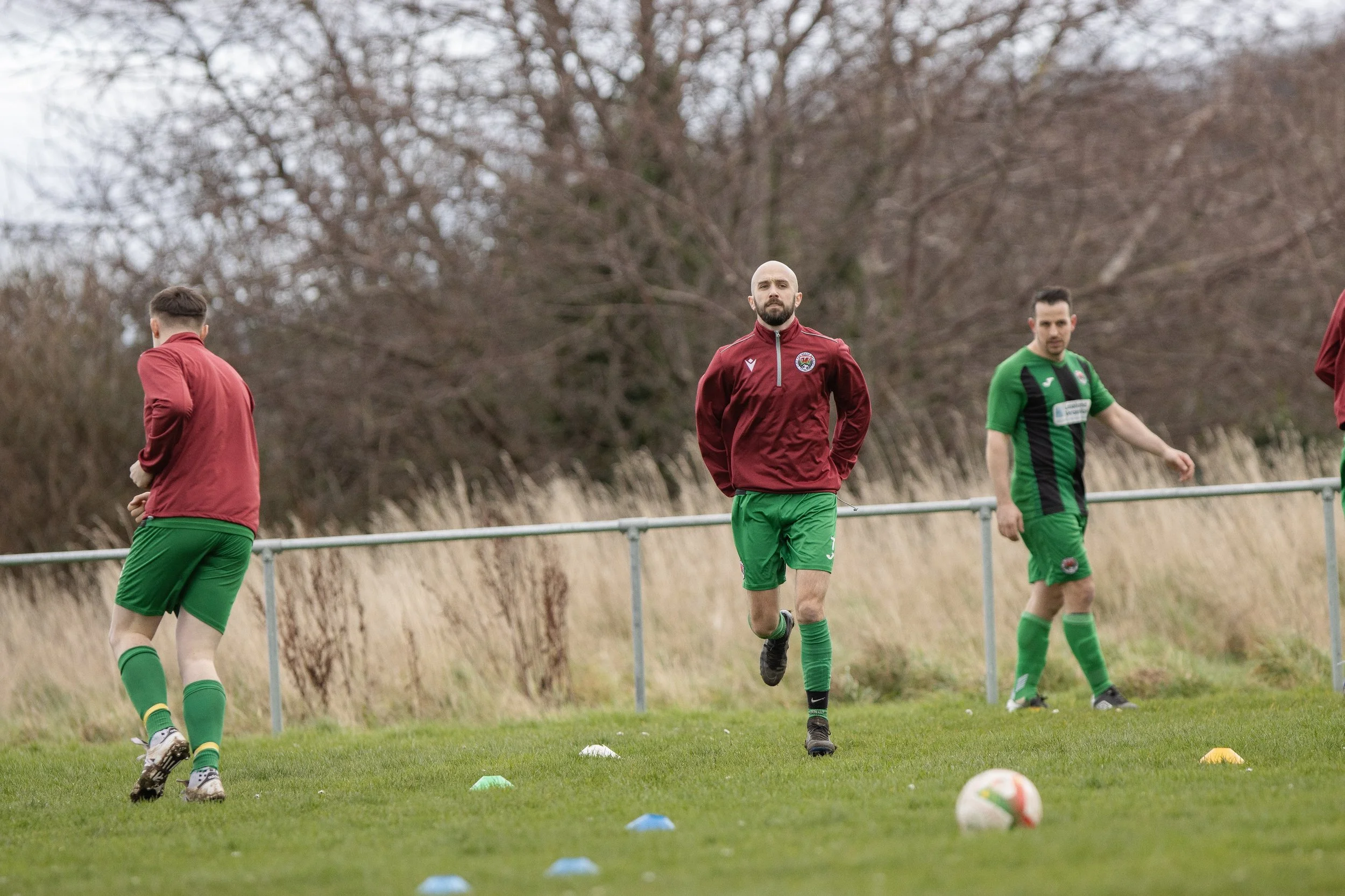 Soccer players practicing on the field, wearing green shorts and maroon jackets, with cones and a soccer ball on the ground.