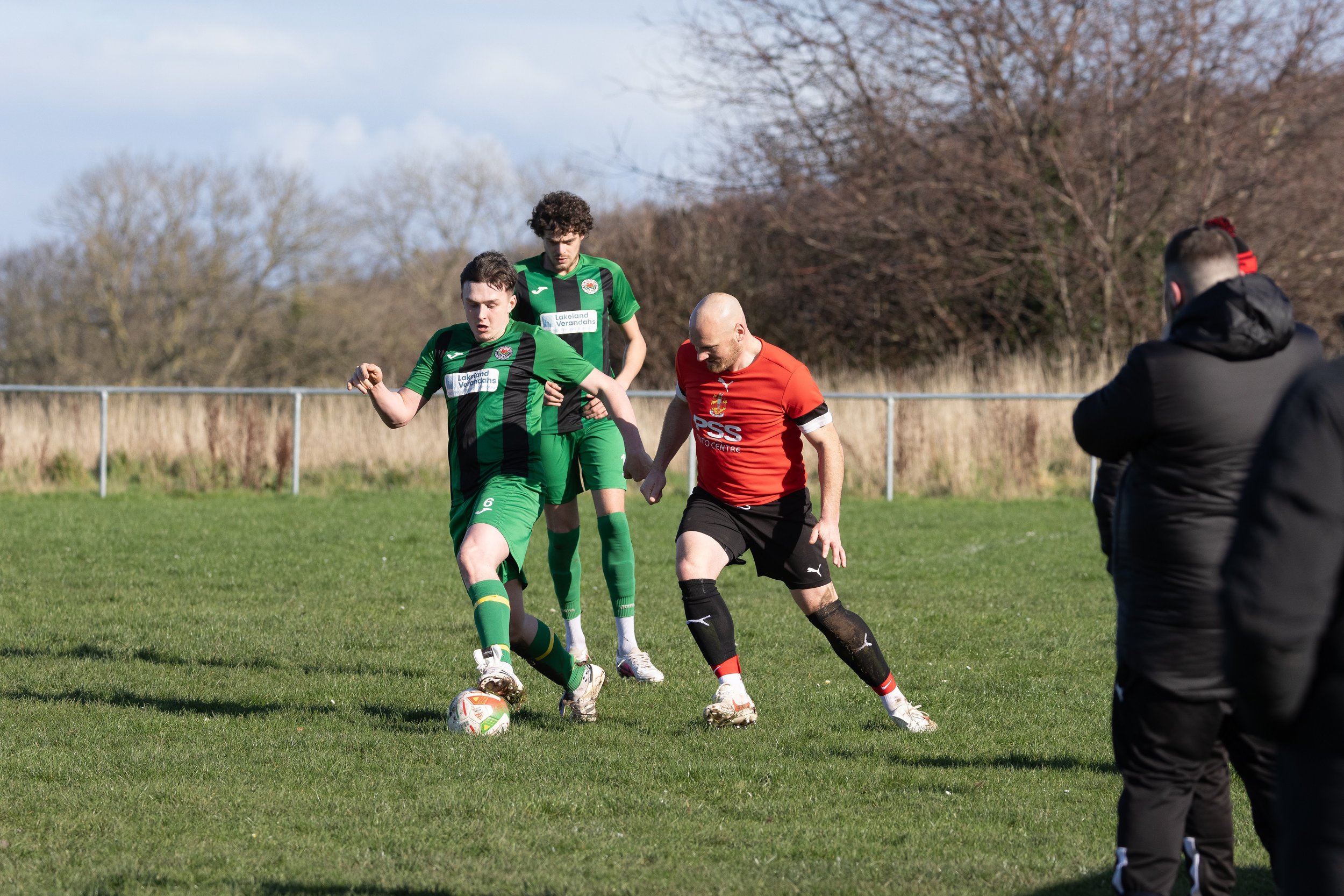 A soccer match with two players in green jersey competing against a player in a red jersey. The player in green is controlling the ball while the red player is attempting to block him. Additional players and spectators are visible in the background a