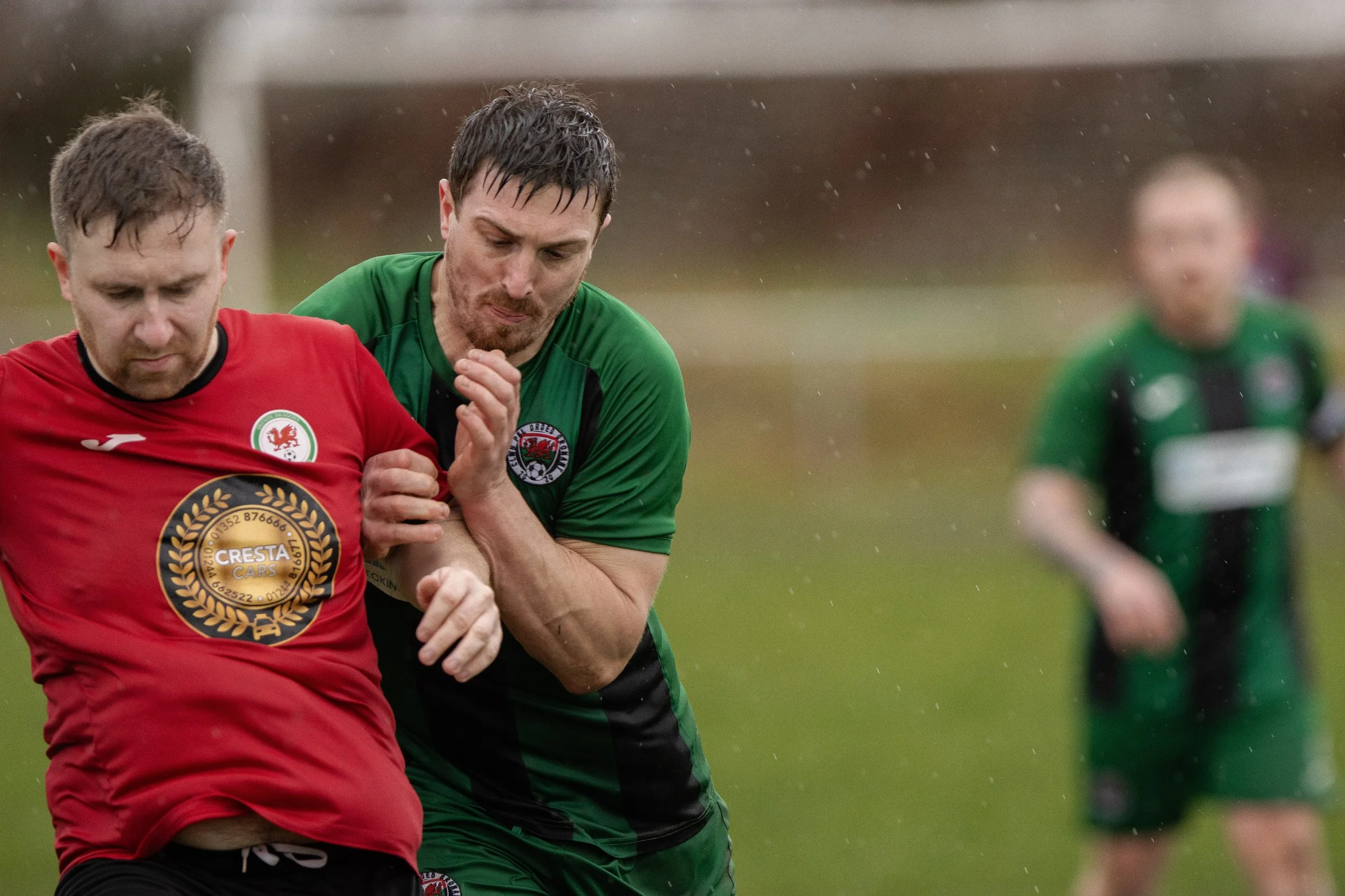 Two soccer players engaged in a match, one wearing a red jersey and the other in a green jersey, competing for possession of the ball on a rainy day.