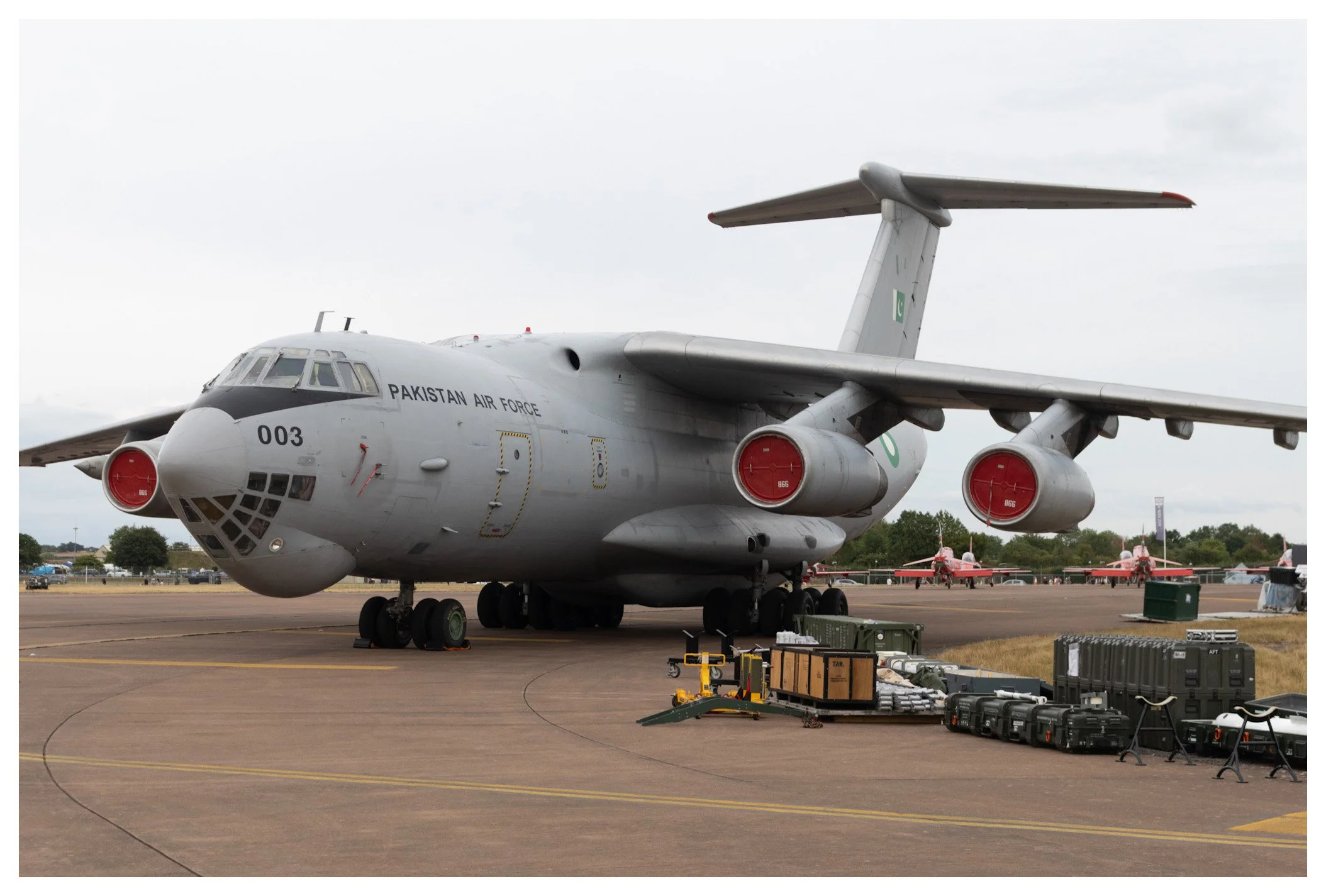 A Pakistan Air Force military aircraft parked on an airfield, with equipment and other aircraft in the background.