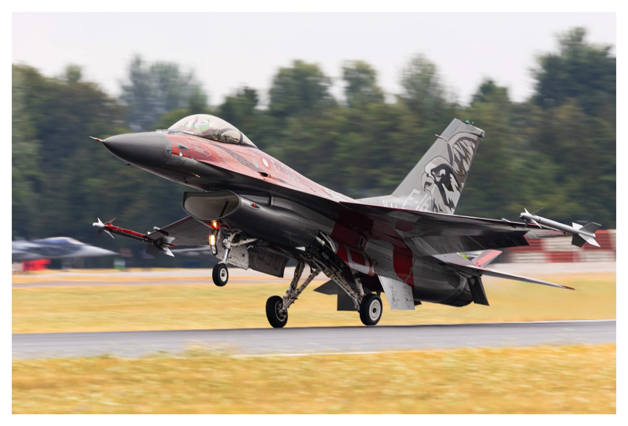 F-16 fighter jet taking off from runway, with blurred background of trees and airport facilities.