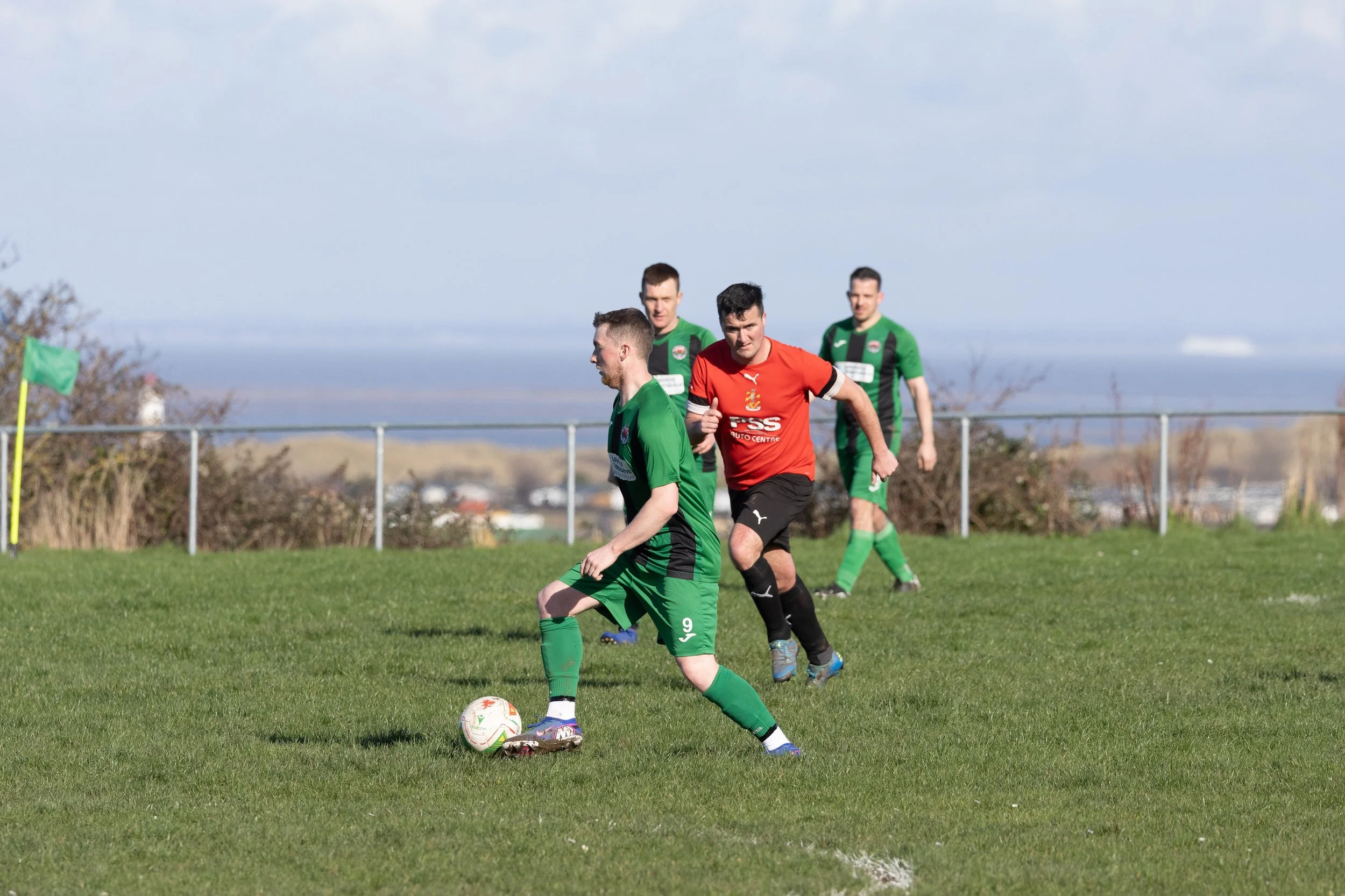 Four soccer players on a grassy field, three in green jerseys and one in red, with the player in green controlling the ball.