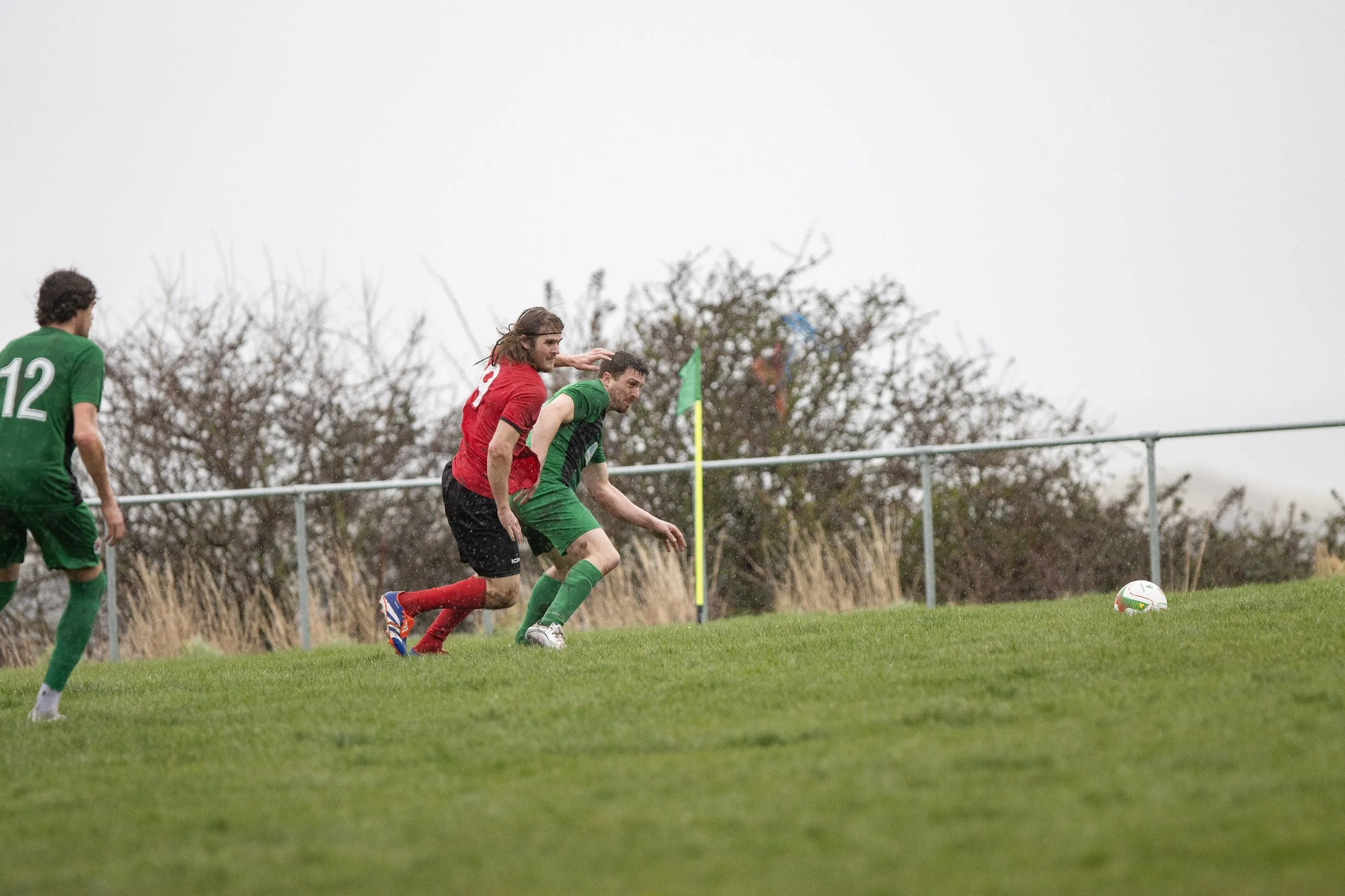 Soccer players in green and red jerseys compete for the ball near the corner flag on a grassy field, with cloudy sky in the background.
