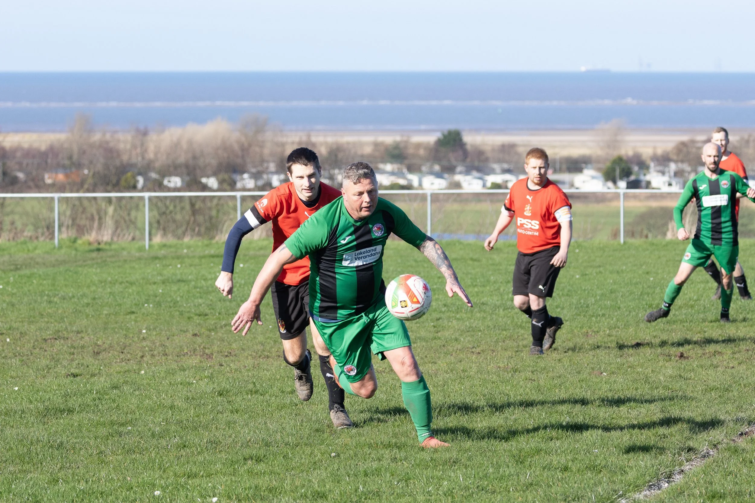 Soccer players in green and red jerseys chasing a ball on a grassy field with a body of water and distant landscape in the background.