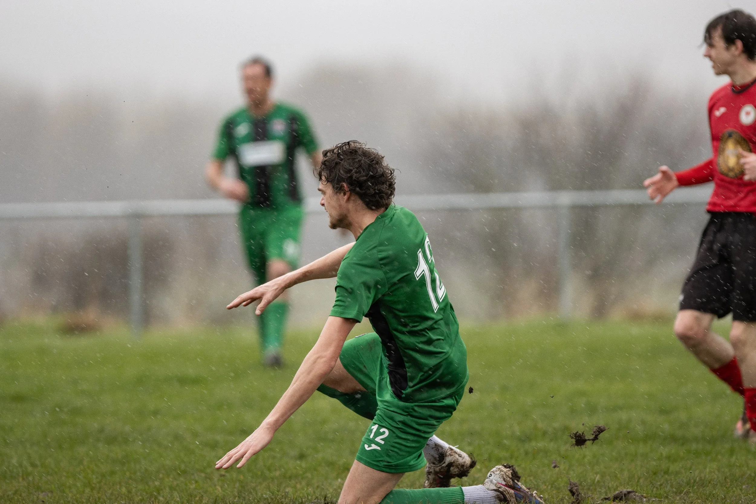 Soccer player wearing a green uniform with the number 12 falling on a wet grass field during a game, with two other players and a coach or referee in the background.