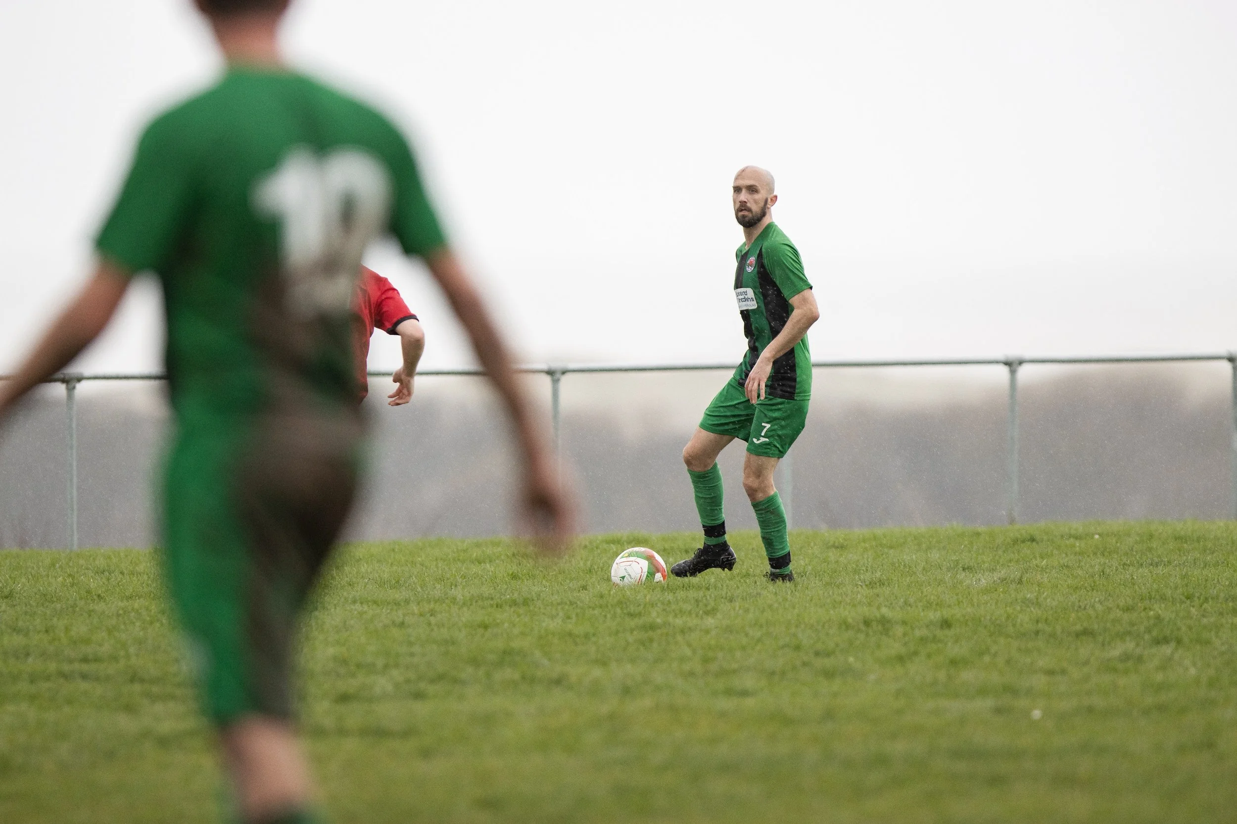 A soccer player stands on the field with a ball at his feet, wearing a green uniform with the number 7 on his shorts. Other players are visible, one blurred in the foreground and another partially seen in the background, in a game setting on a grassy