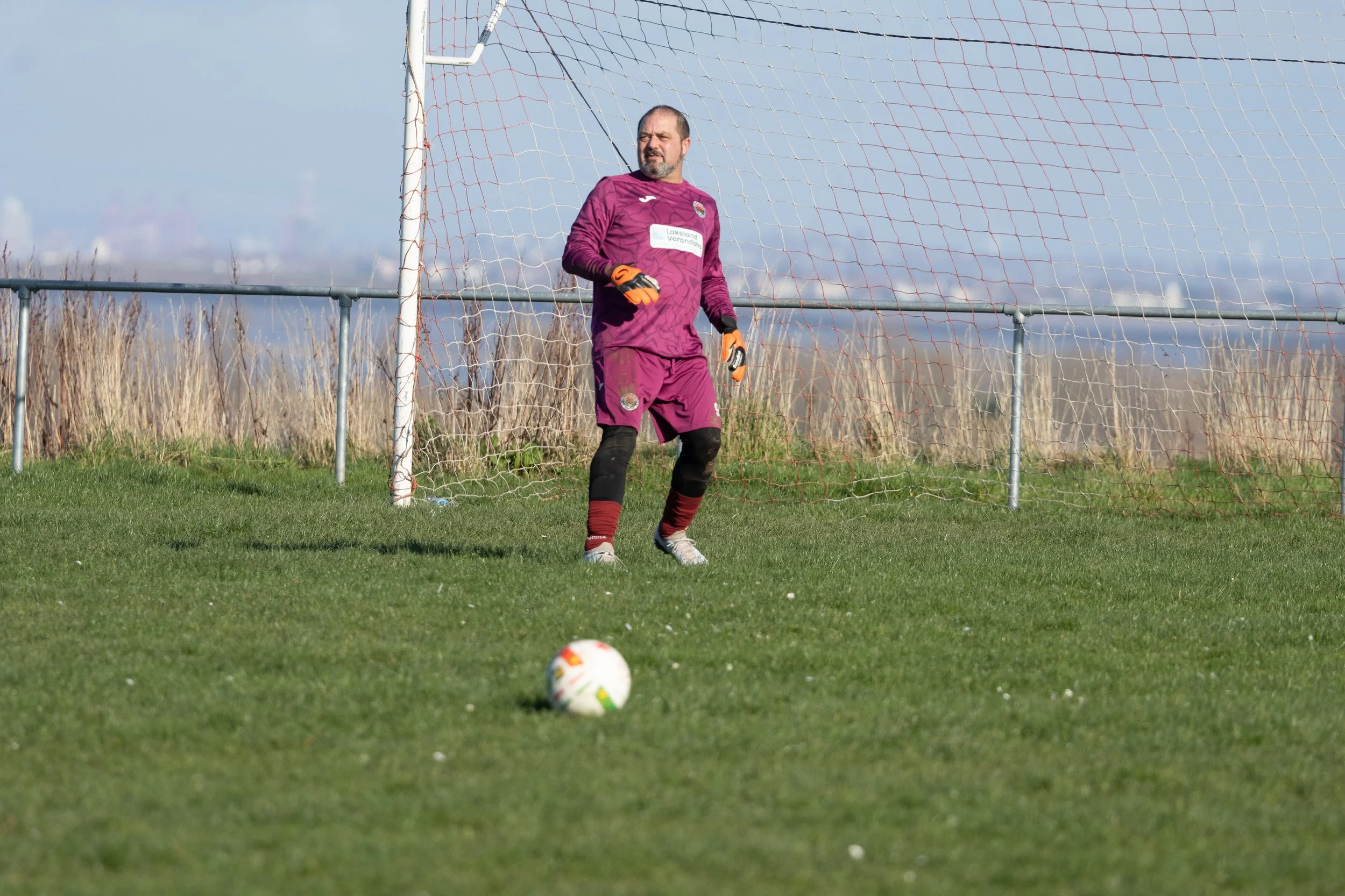 A soccer goalkeeper standing on the field in front of the goal, with a soccer ball on the grass in front of him, during daytime with a cloudy sky.