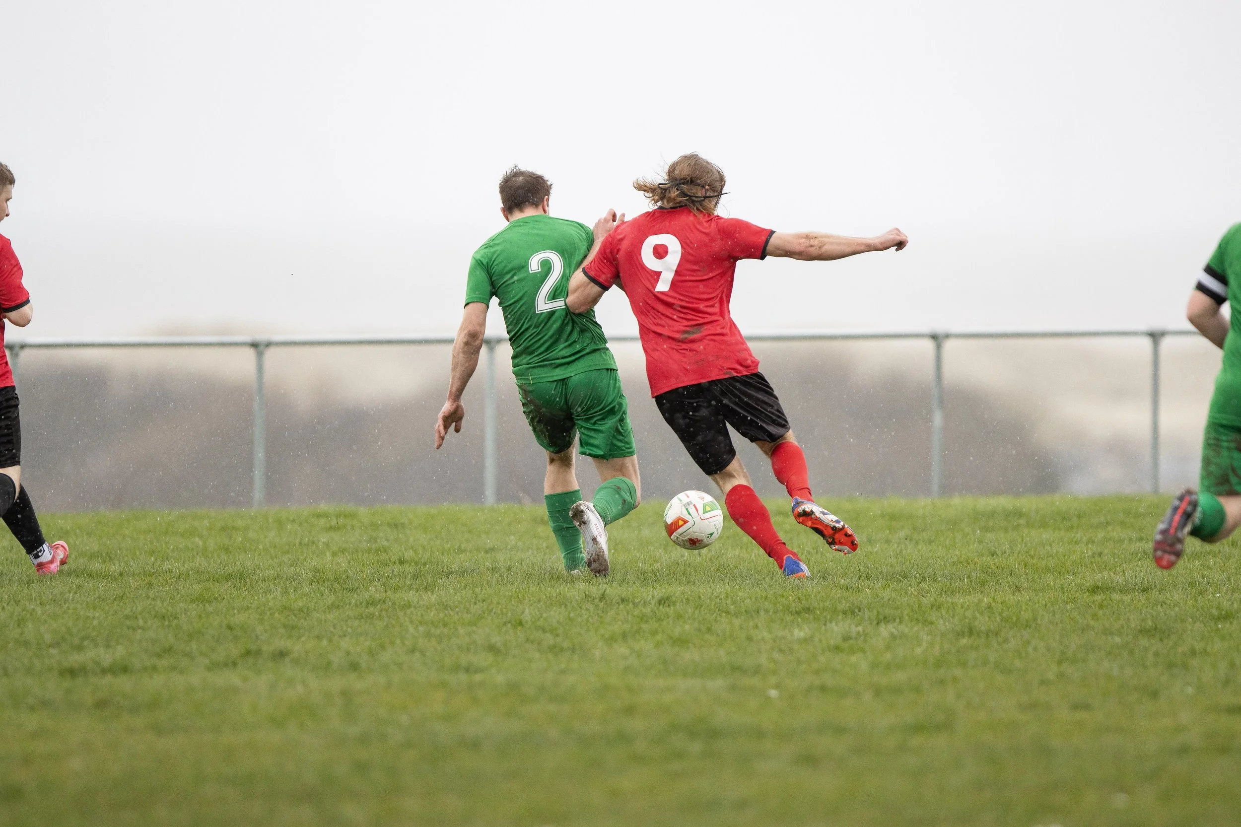 Two soccer players, one in a green jersey with number 2 and the other in a red jersey with number 9, are competing for the ball on a grassy field. The player in red is kicking the ball while the player in green is closely defending. Other players are