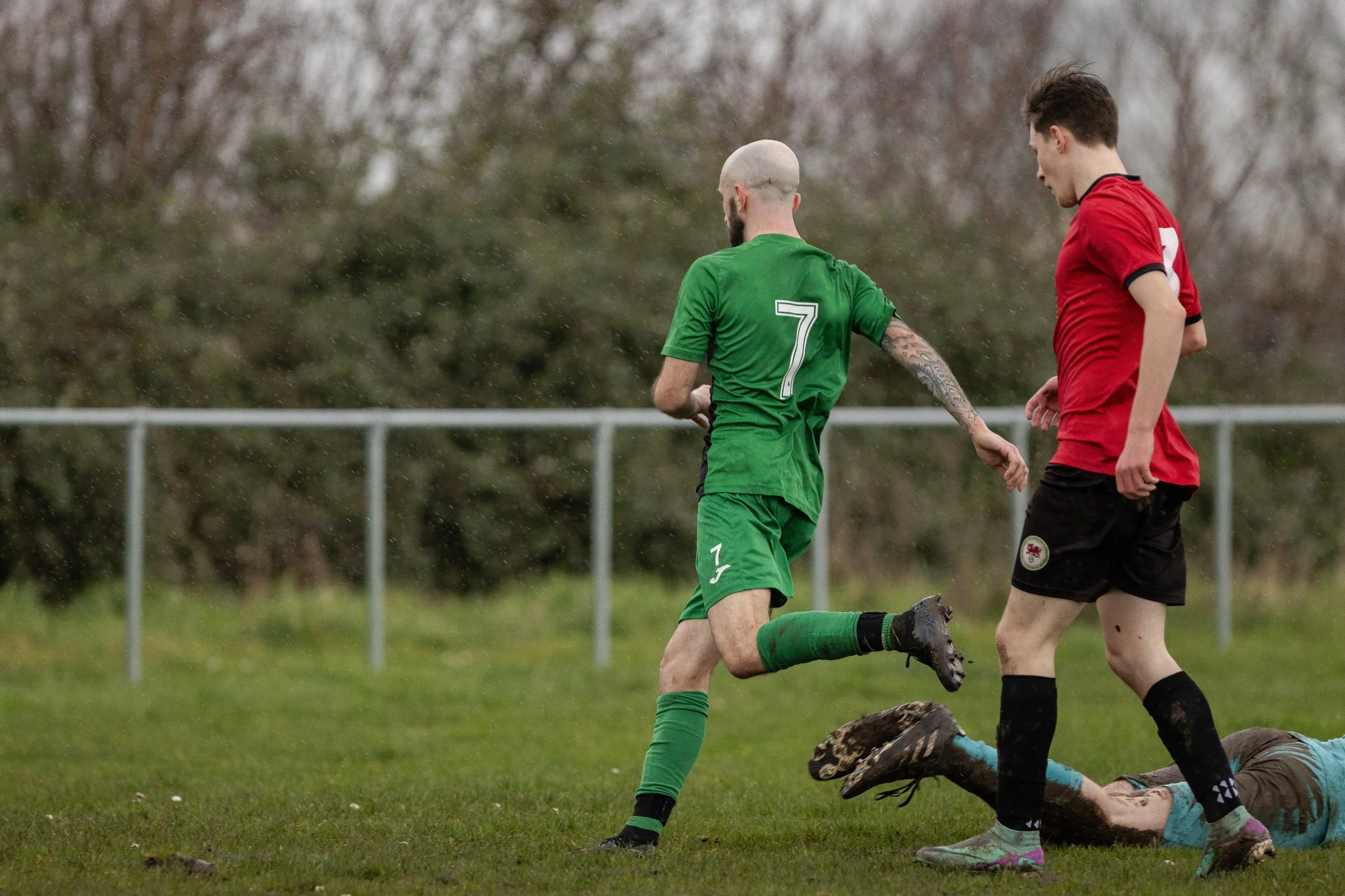 Two soccer players, one in a green uniform with the number 7, and the other in a red uniform, are on a muddy field. The player in green appears to be running away from a fallen player dressed in blue, who is on the ground, over the muddy grass.