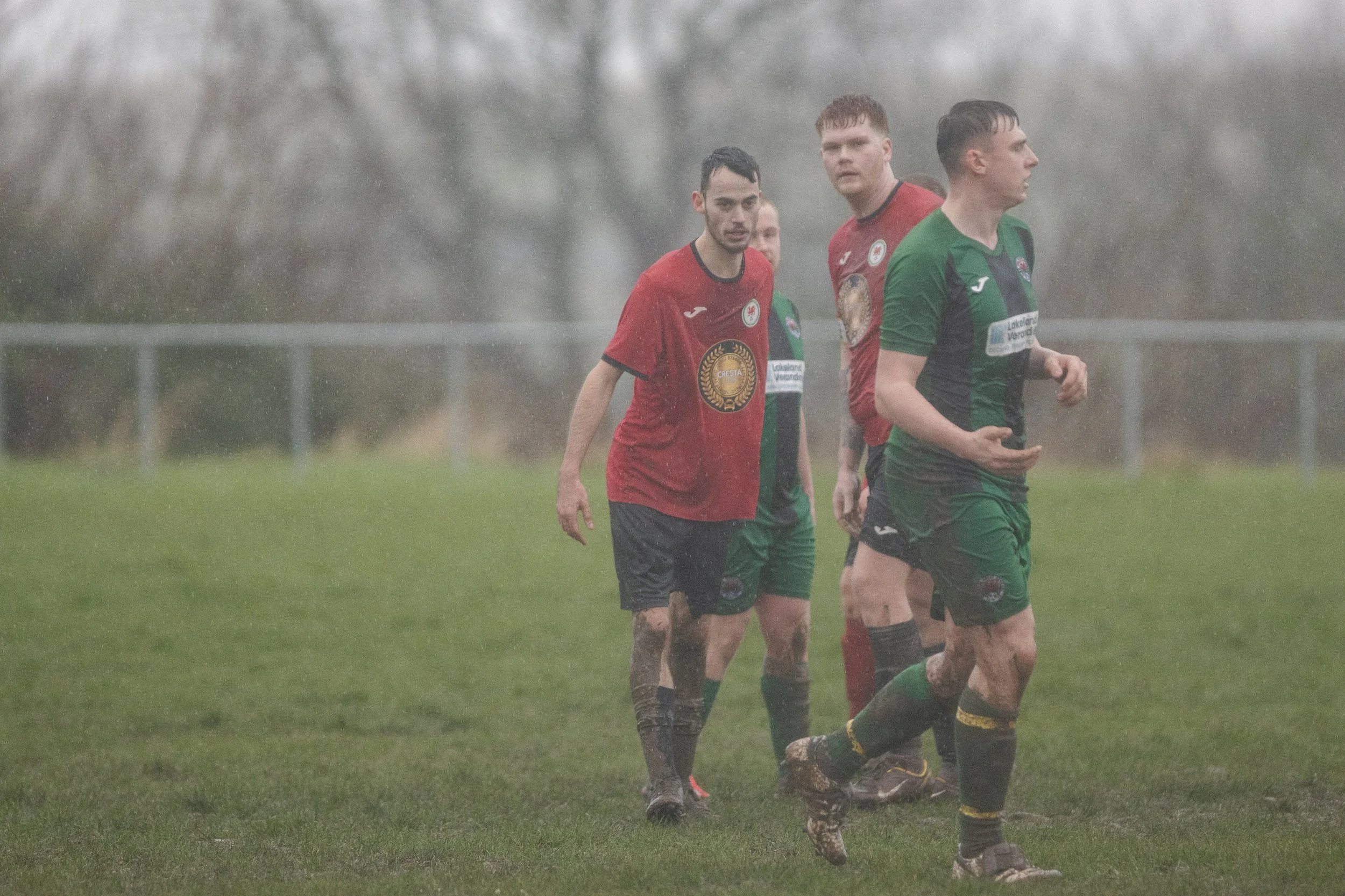 Four soccer players walking on a wet, muddy field in the rain. Two players are wearing red jerseys and two are in green jerseys.