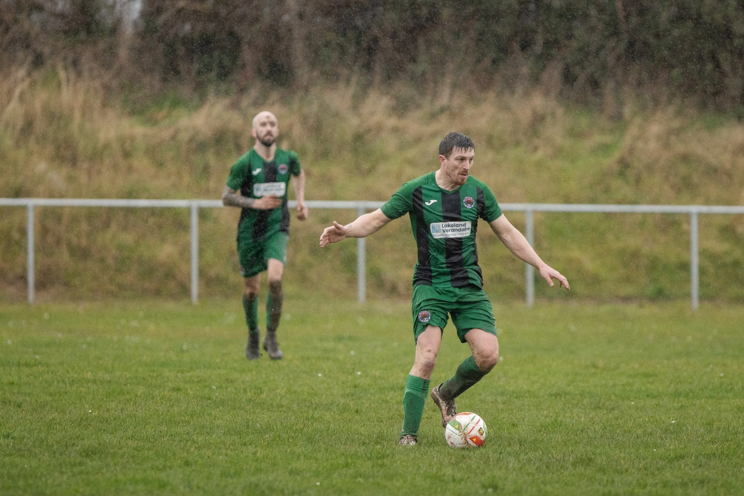 Two soccer players in green and black uniforms on a rainy field, with one preparing to kick a soccer ball and the other running in the background, with grassy hillside and a metal fence in the distance.