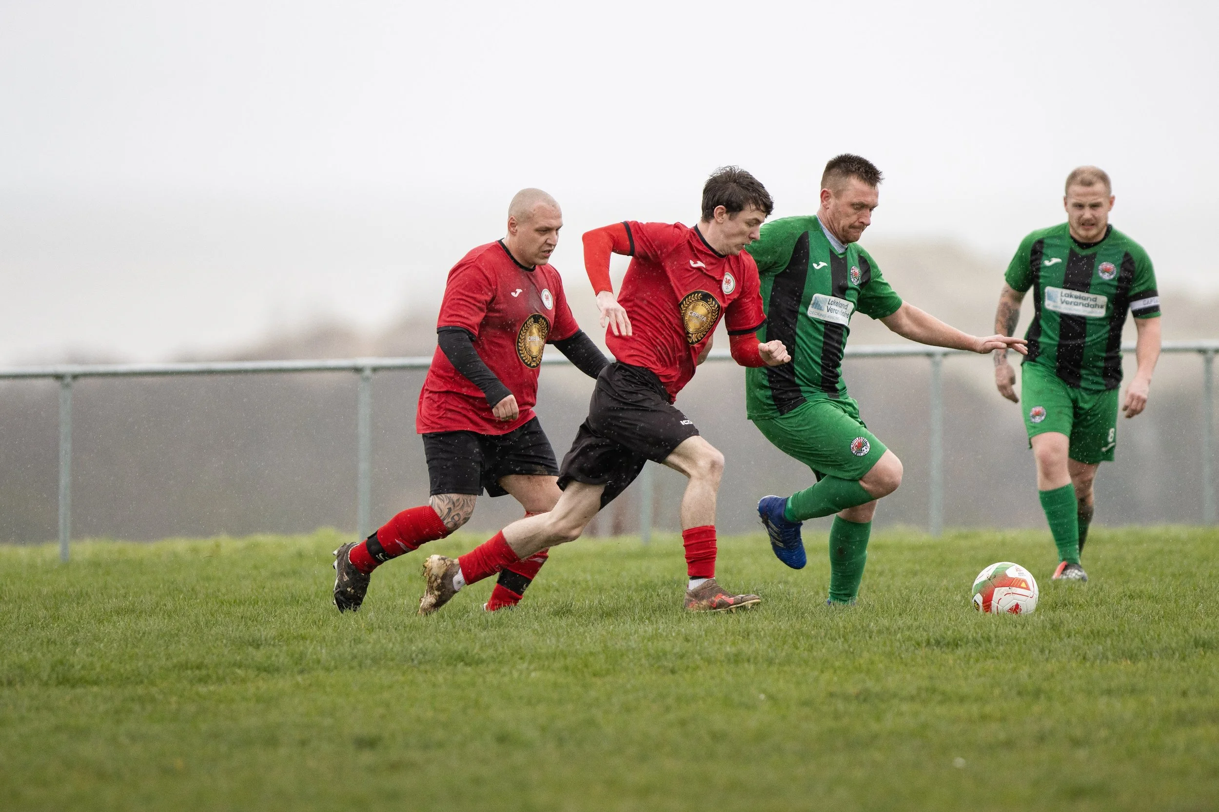 Four soccer players competing for possession of the ball on a grassy field in rainy weather.