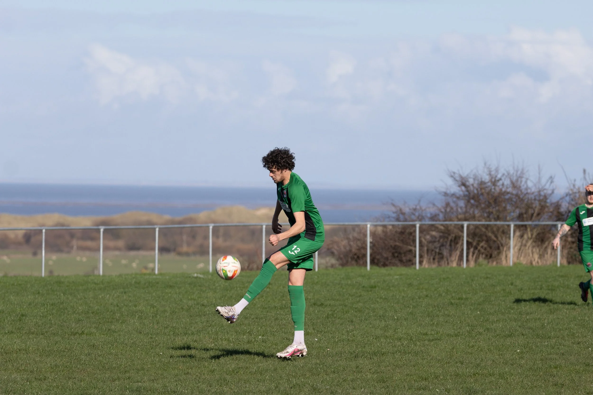 Soccer player in green jersey kicking a soccer ball on a grassy field with a blue sky and distant trees.