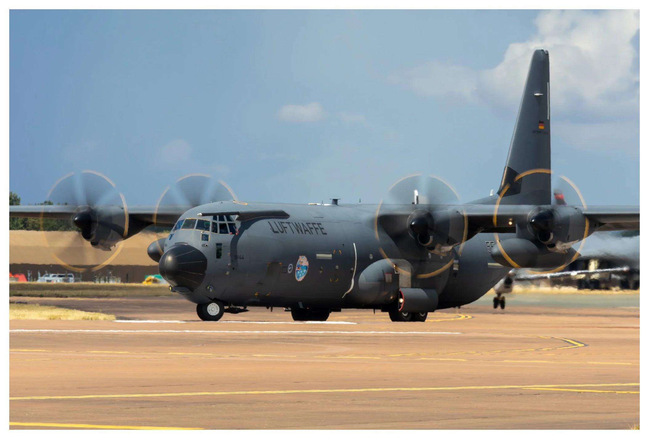 Military cargo airplane on runway, propellers spinning, with blue sky and some clouds in background.