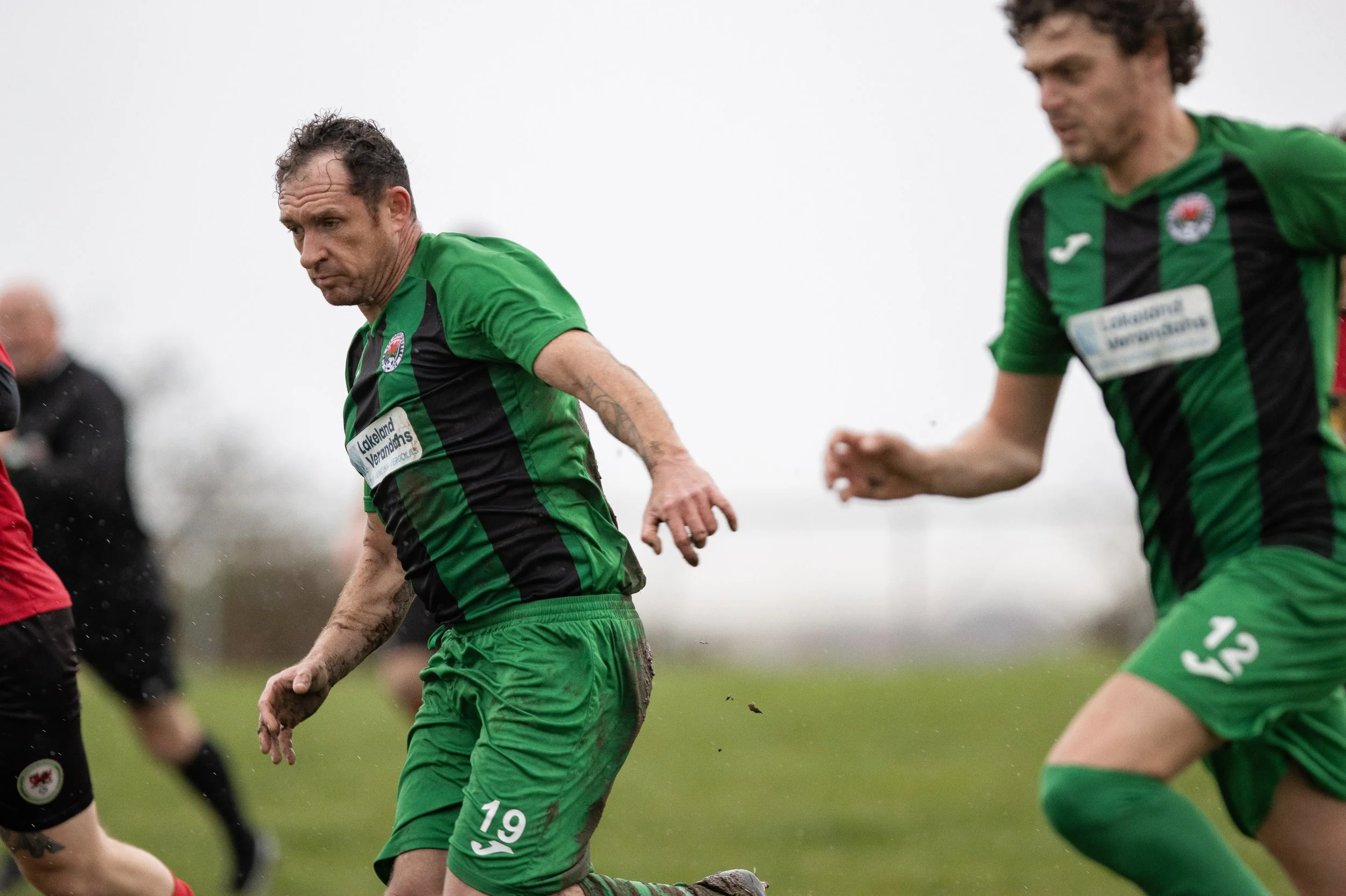 Two soccer players in green jerseys, numbered 19 and 15, playing on a grass field during a match, with a blurred background and overcast sky.