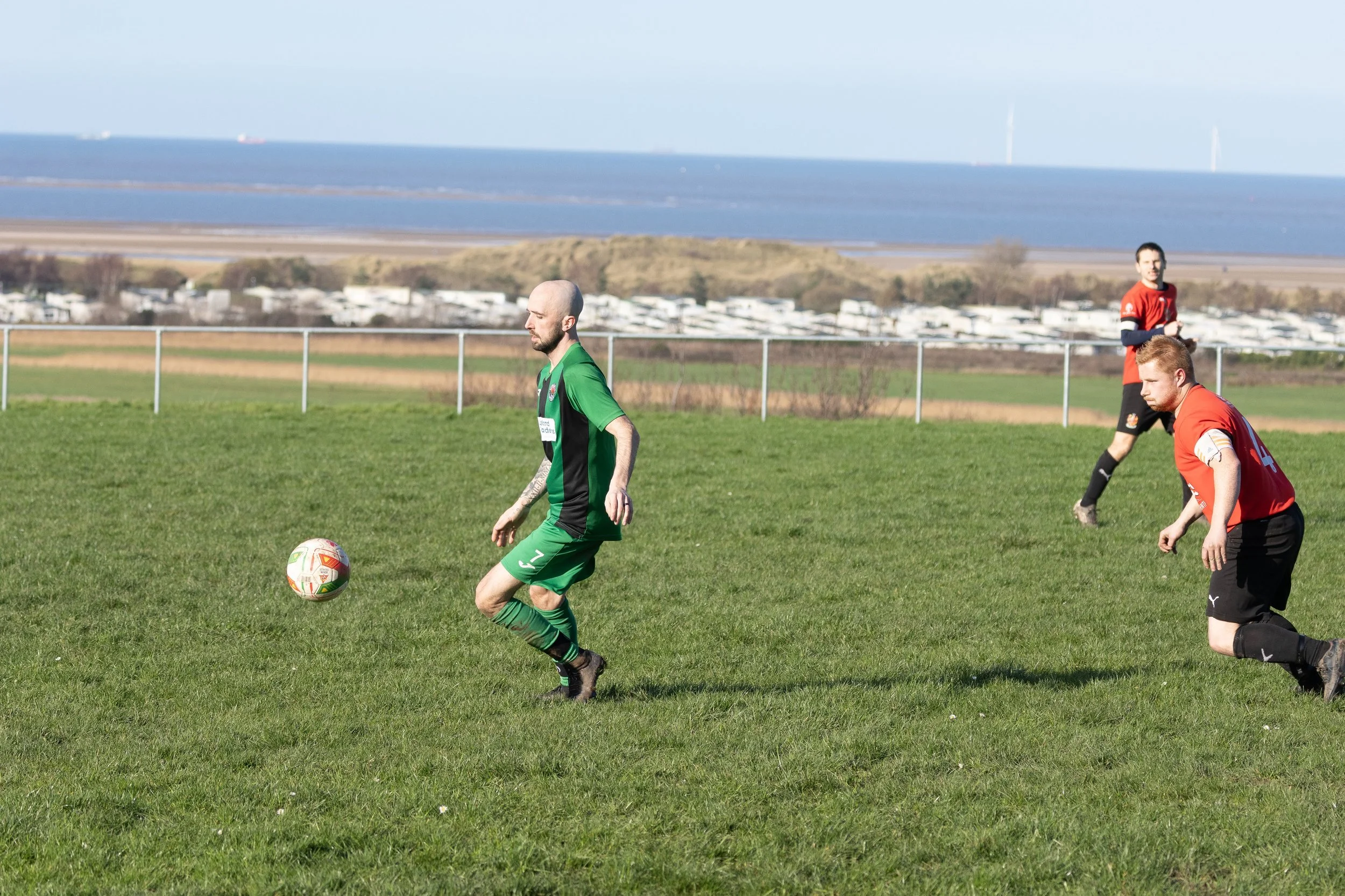 Three men playing soccer on a grassy field with a body of water and wind turbines in the background.