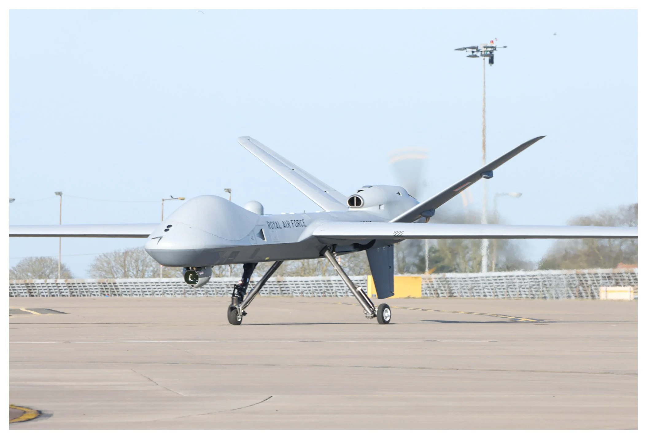 A military drone marked 'Royal Air Force' on a runway with a sky background and an aircraft in the distance.