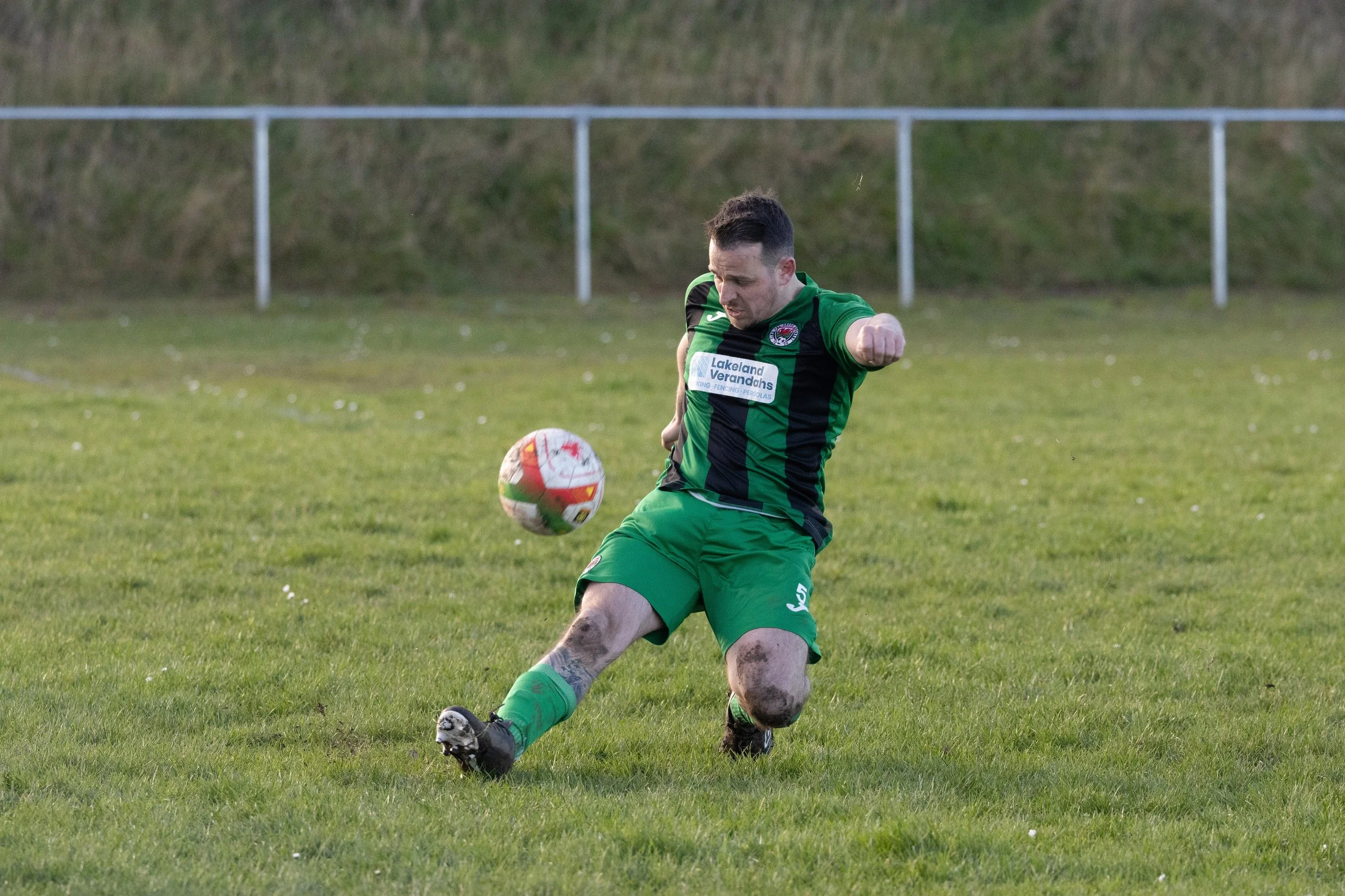 A male soccer player in a green and black uniform kicks a soccer ball on a grassy field during a match, with a metal fence and hill in the background.