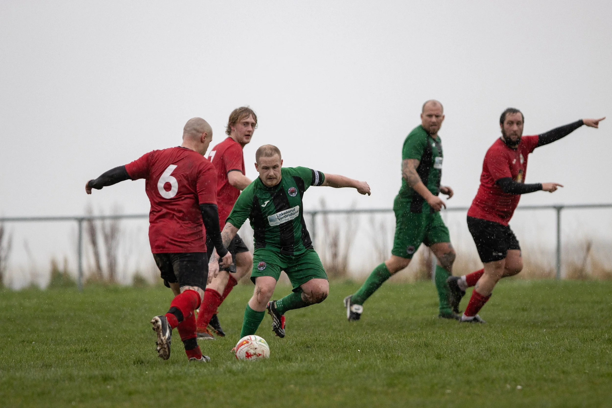 Soccer players competing on a rainy field, with some players in red and others in green jerseys, focusing on a player in green who is about to kick the ball.