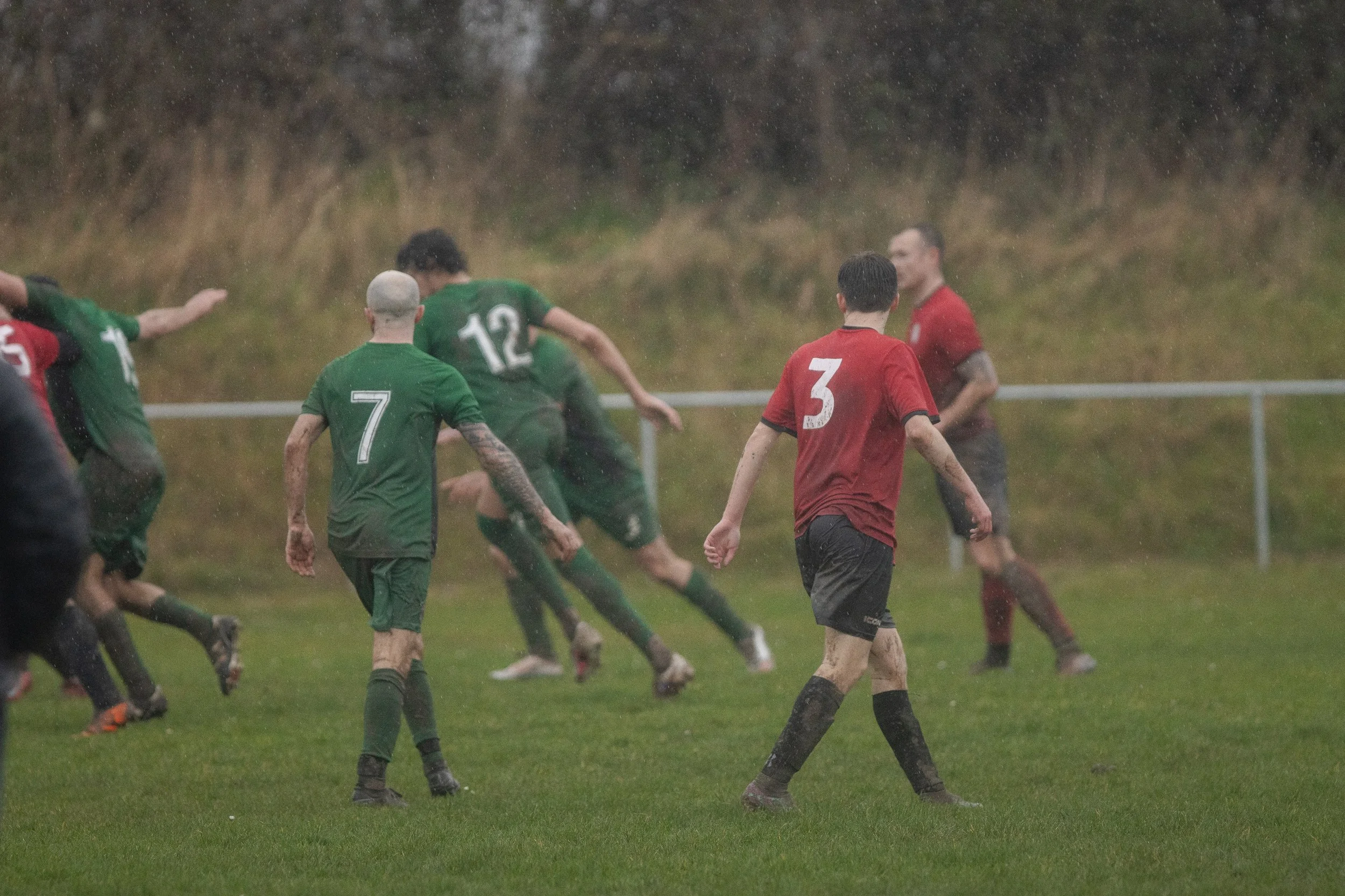 Soccer players on a muddy field during a rainy match, wearing green and red jerseys.