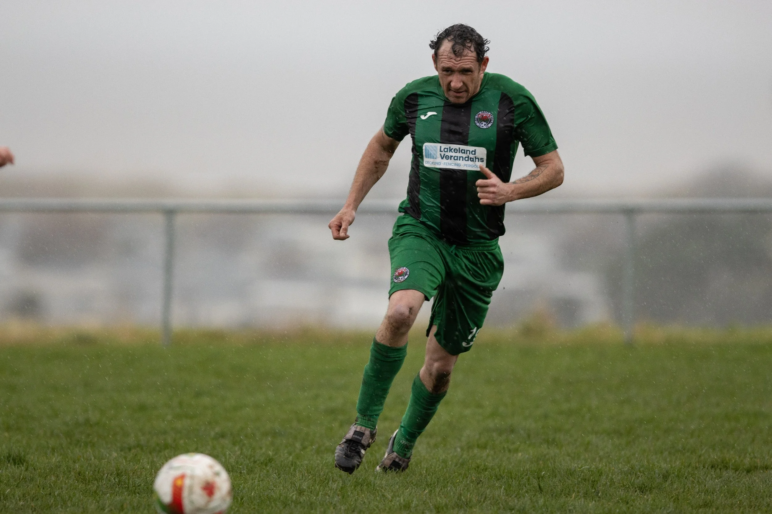 A soccer player in green and black uniform running on a wet field with a ball in front of him on a rainy day.