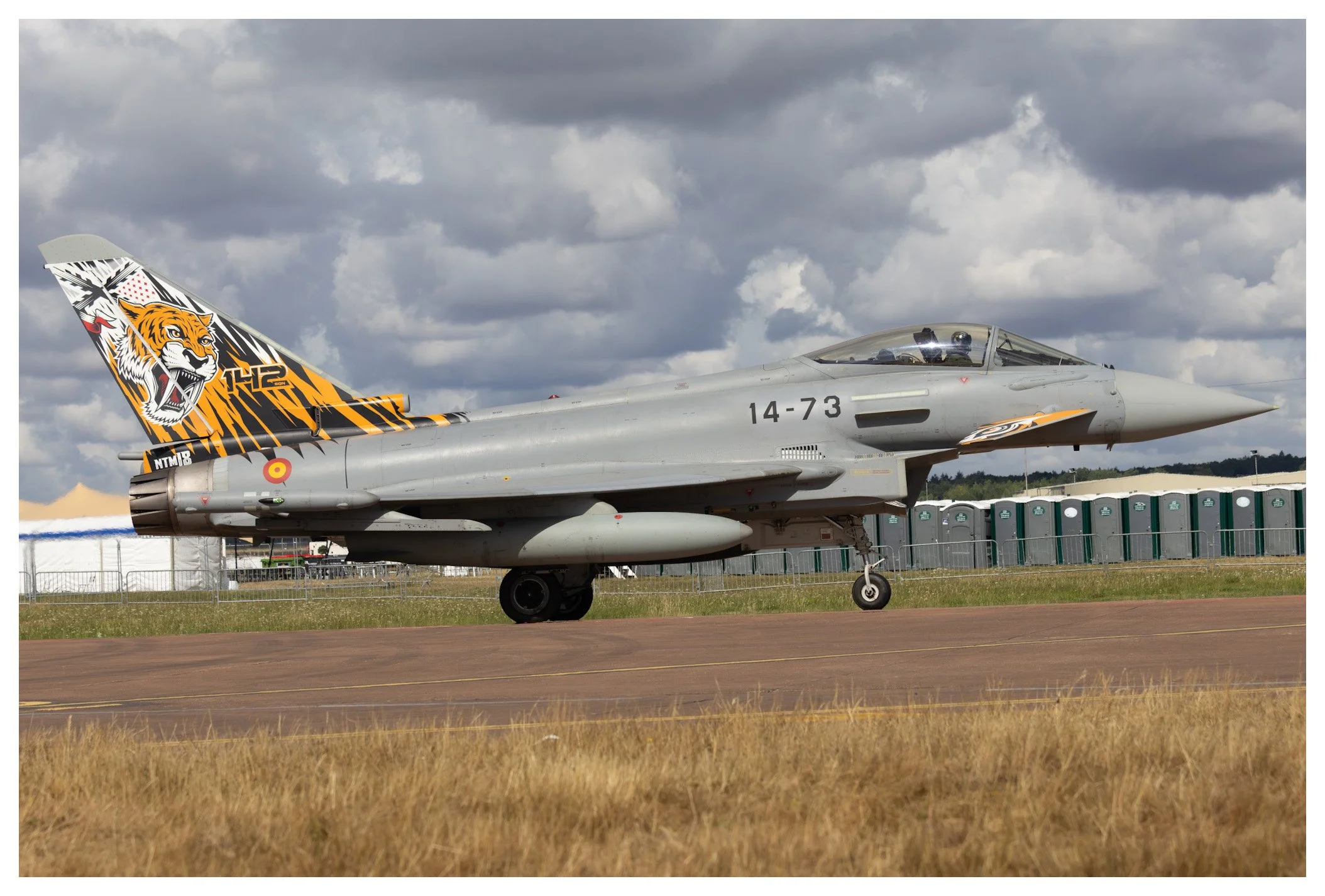 A fighter jet with a tiger logo on the tail parked on a runway, with portable toilets and a cloudy sky in the background.