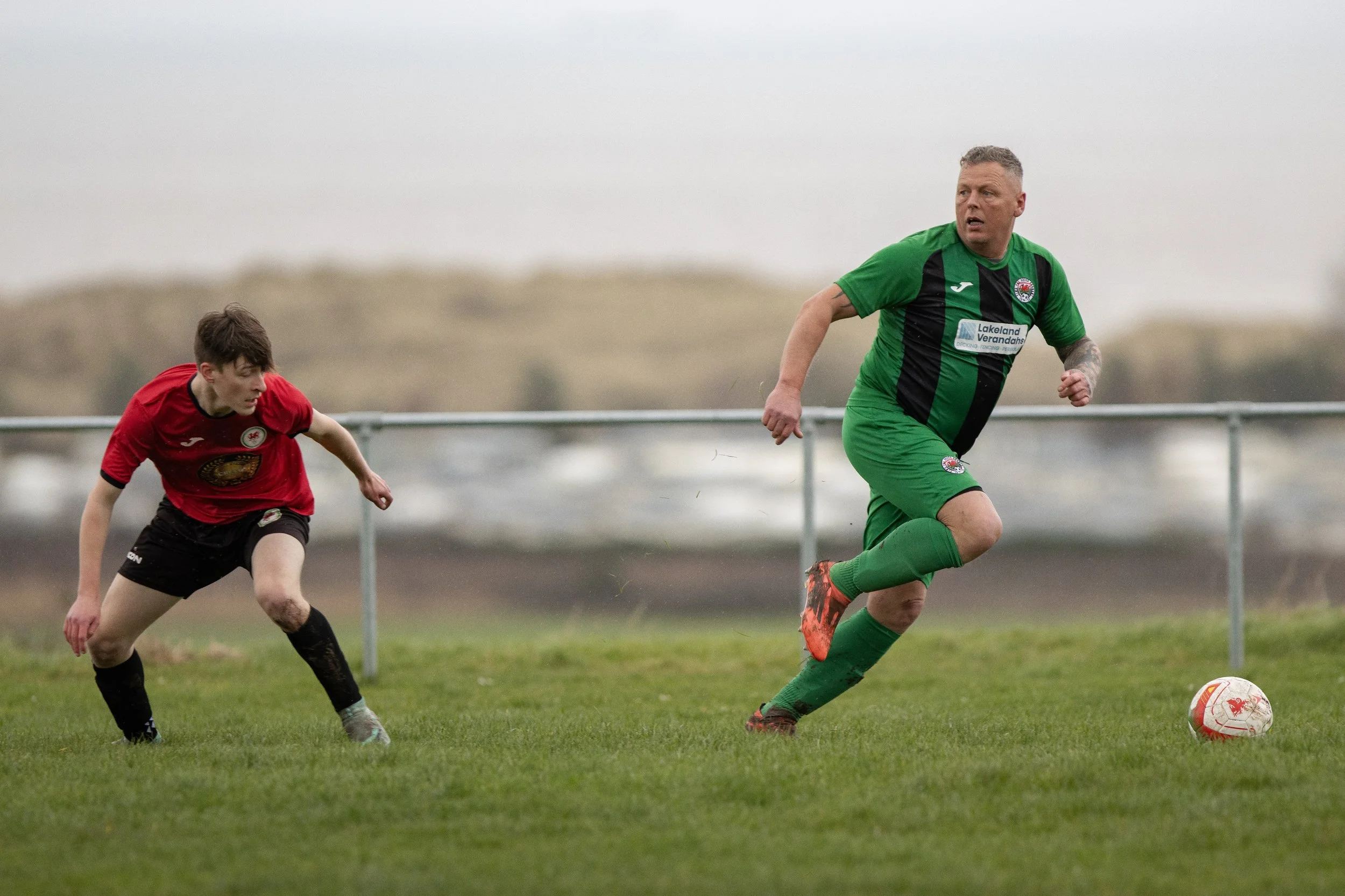 Two soccer players on a grassy field, one in a green and black uniform running towards the soccer ball, the other in a red and black uniform chasing behind.