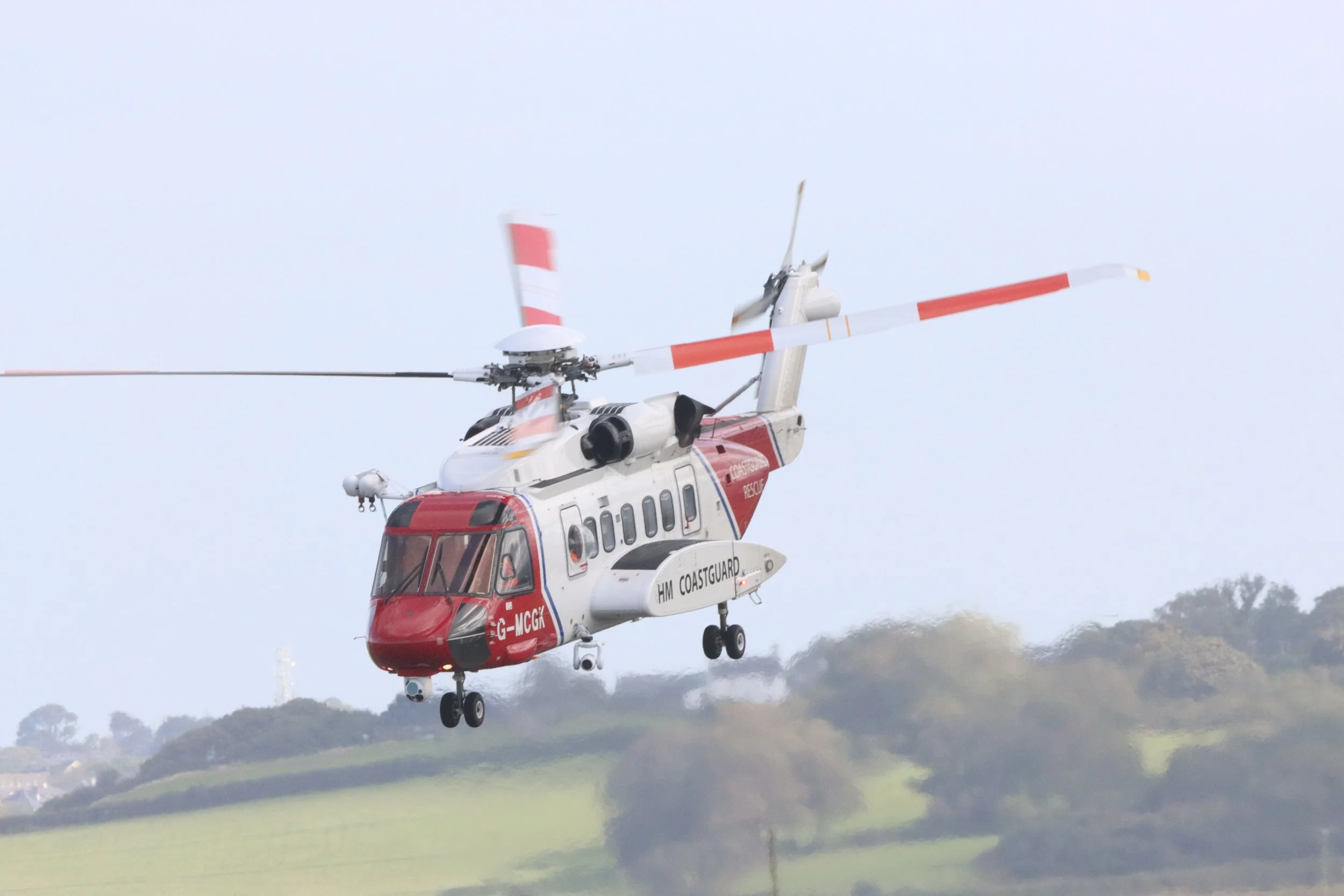 A rescue helicopter flying above a hilly landscape with trees in the background.