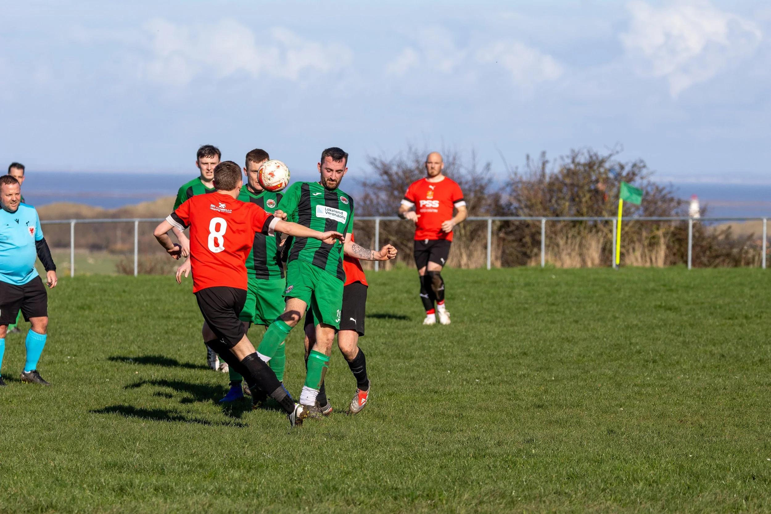 Soccer players on the field competing for the ball during a match on a sunny day, with a scenic background of trees and water.