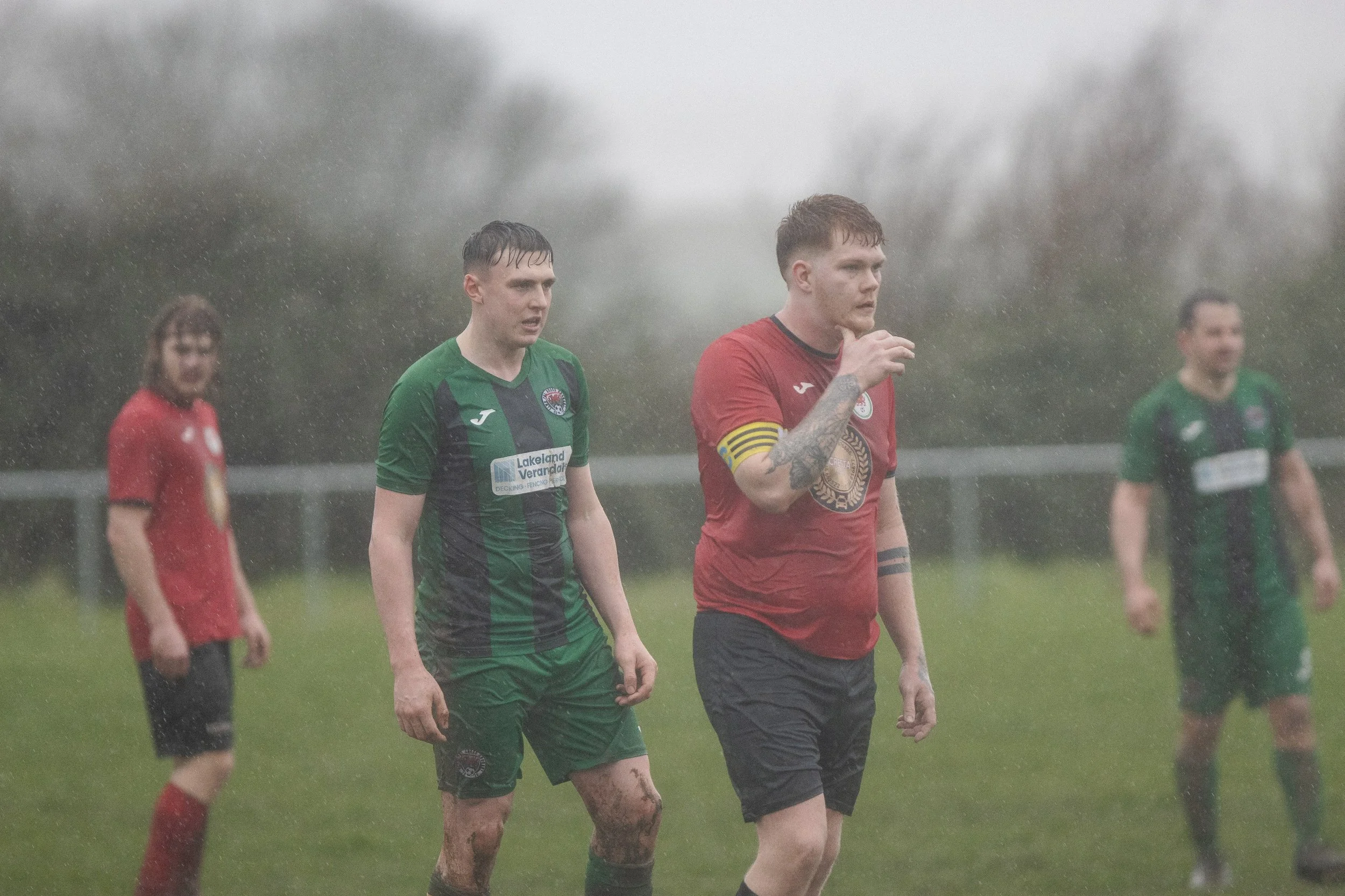 Soccer players on a rain-soaked field, two in green jerseys and one in a red jersey with a yellow captain's armband.