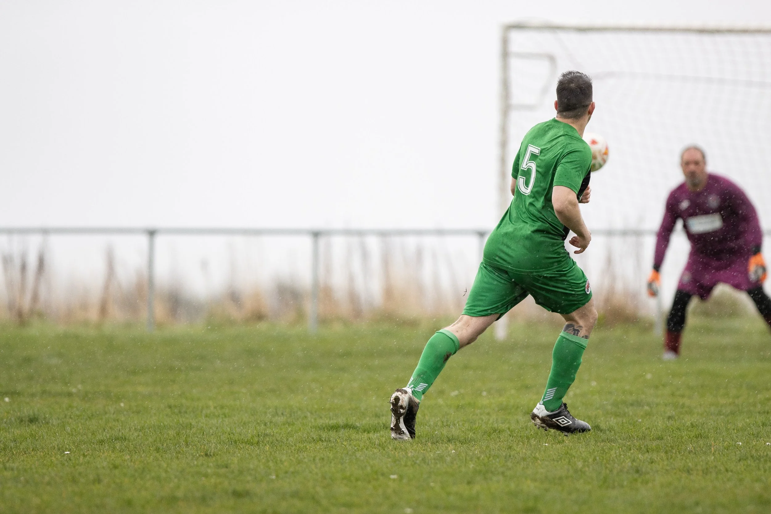 Soccer player in green jersey and shorts with the number 5, running on wet grass towards the goal as goalkeeper in purple jersey and gloves prepares to defend.