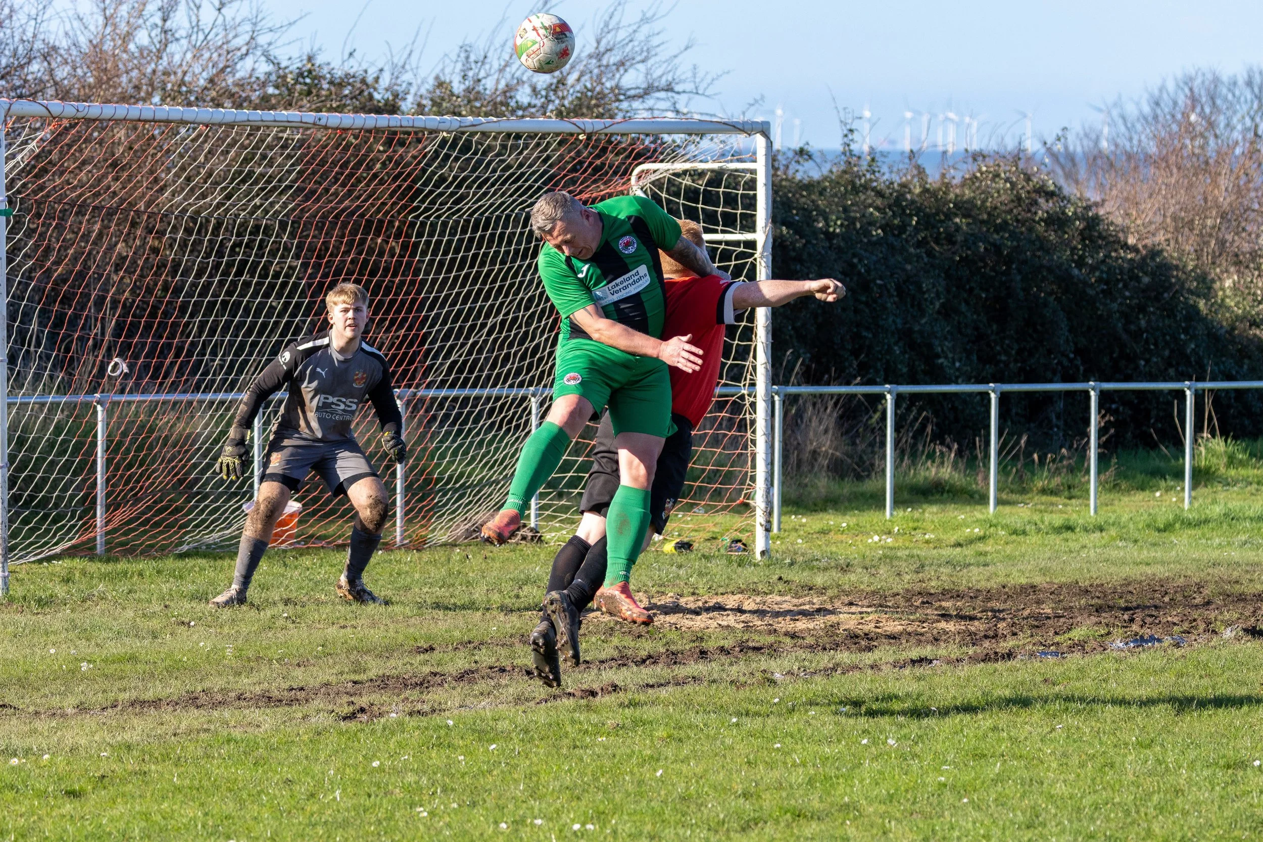 A soccer match with a goalkeeper in black standing near the goal, two players in green and red jerseys contesting an airborne ball, on a grassy field under a clear sky.