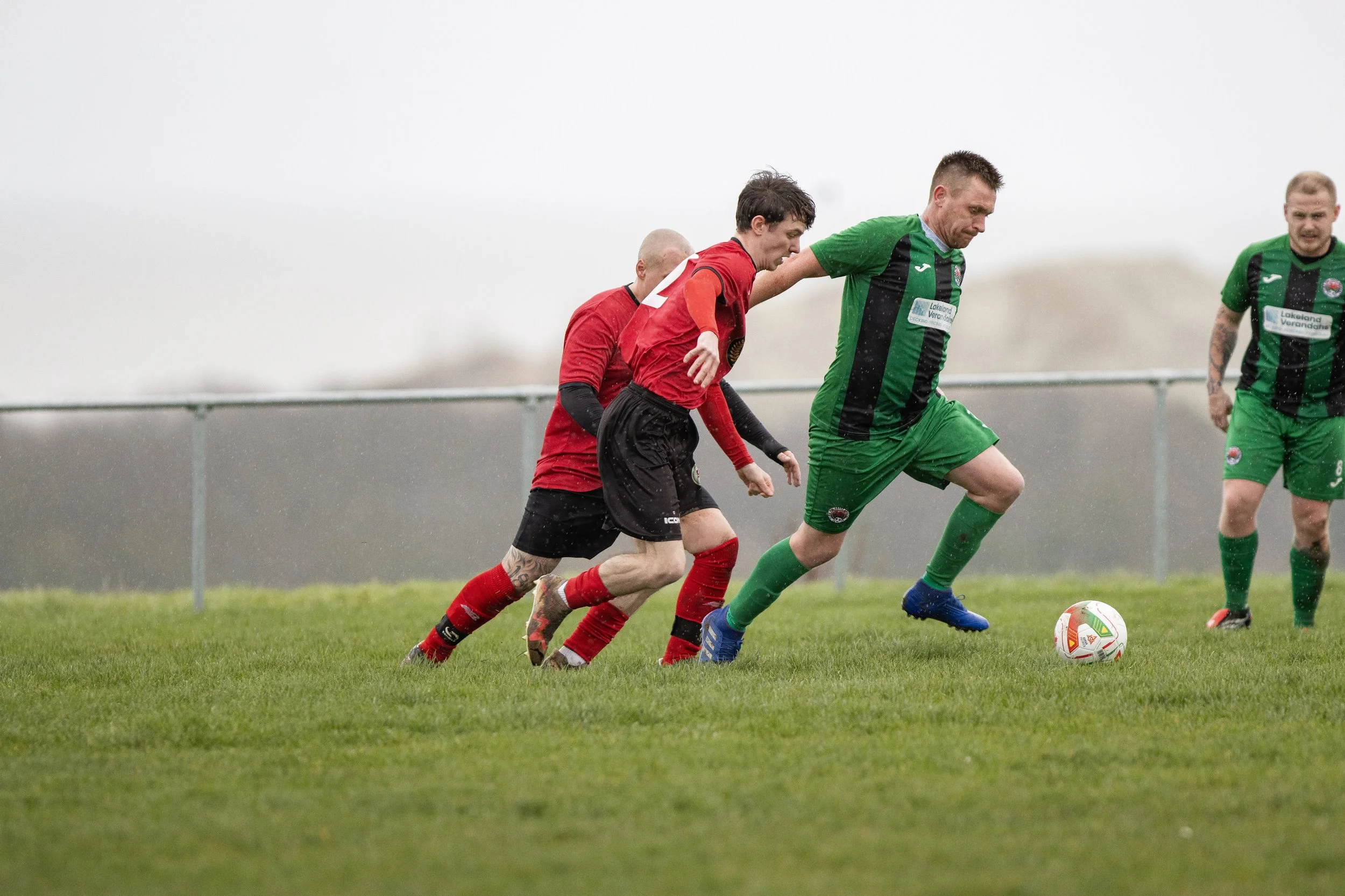 Four soccer players compete for the ball on a rainy field with a gray sky and a fence in the background.
