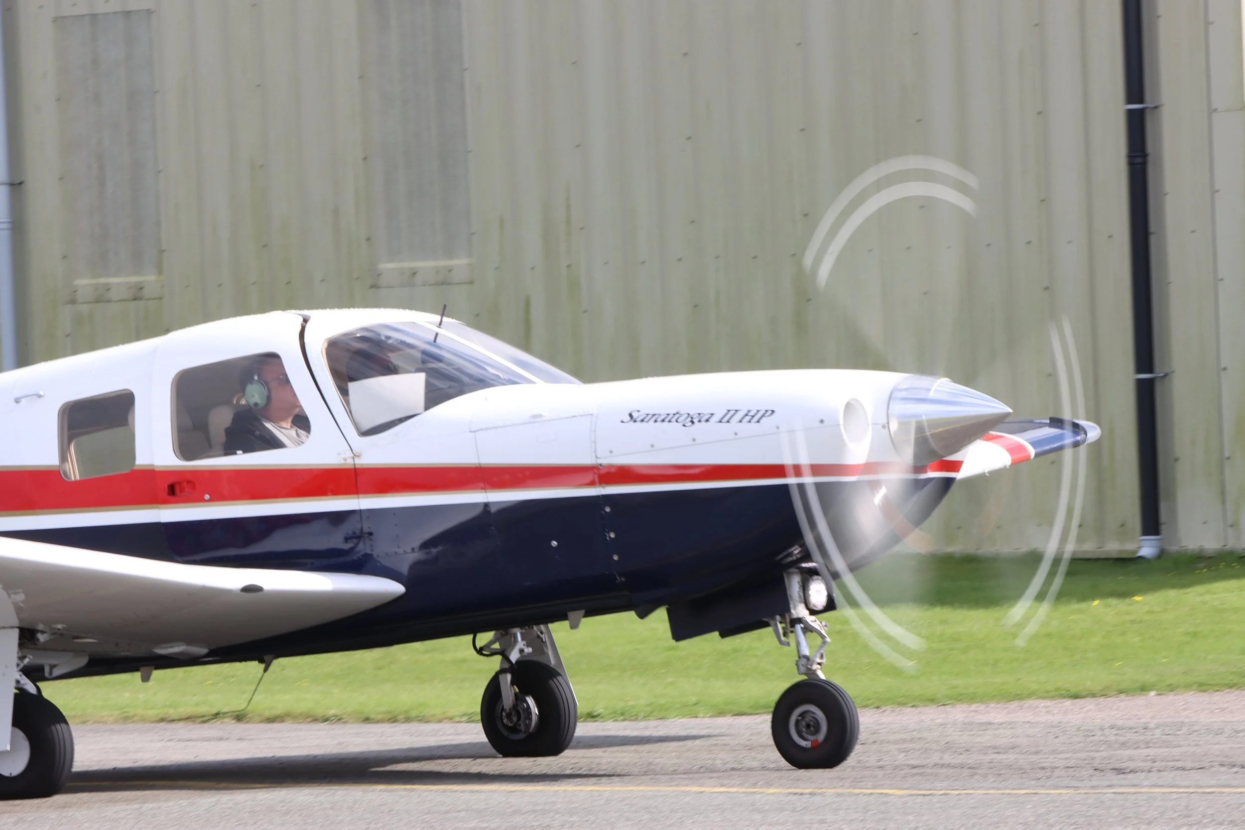 A small aircraft with a white and blue body and red stripe taxiing on a runway, with the pilot wearing headphones visible in the cockpit.
