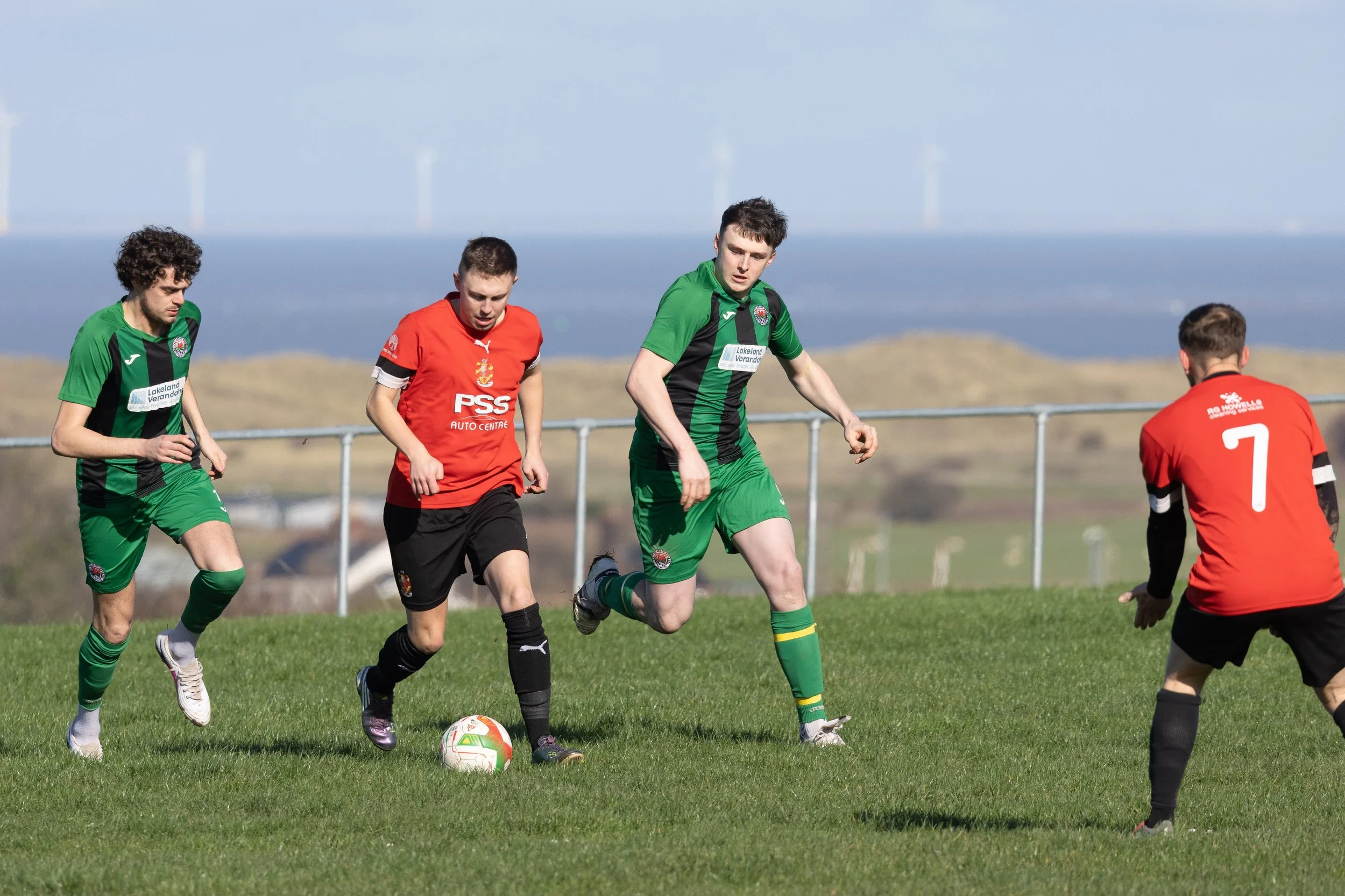 Four soccer players on a grassy field during a match, with two players in green and two in red jerseys, and a soccer ball on the ground.