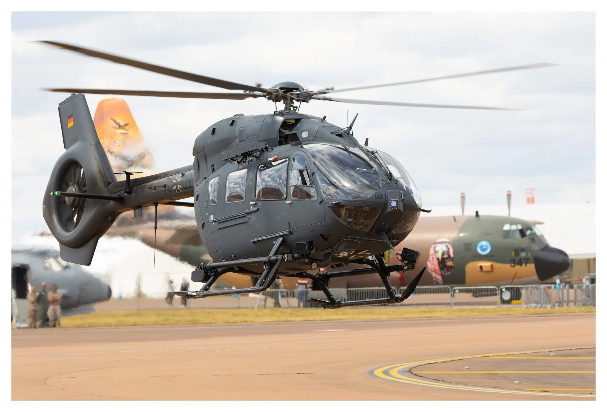 A military helicopter taking off from an airfield, with another aircraft and onlookers in the background.