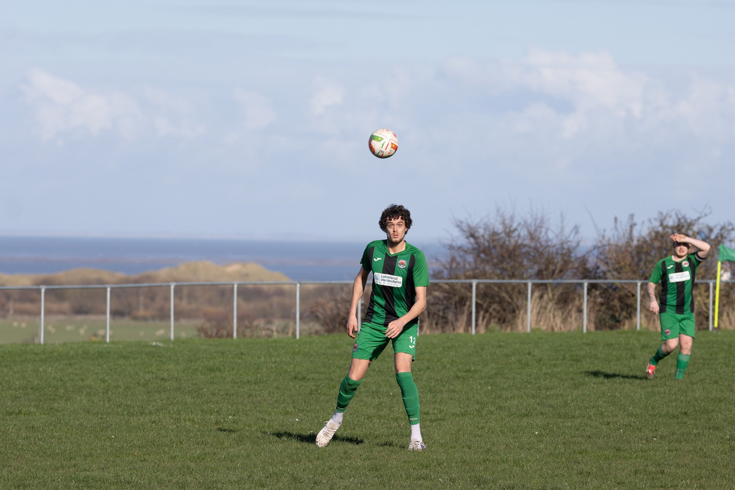 A soccer player in a green and black uniform is about to head the ball on a grassy field with another player in the background.
