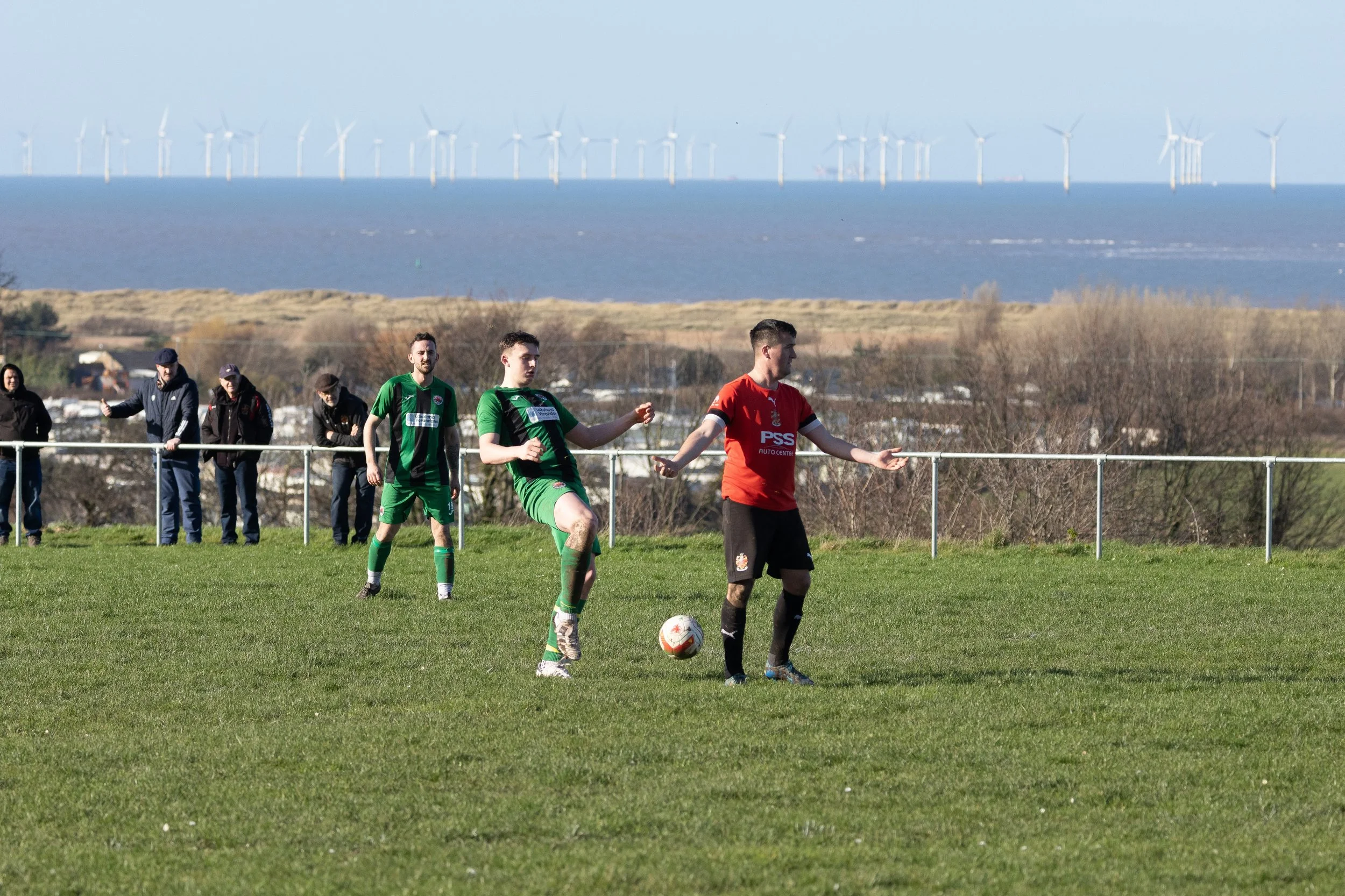 Soccer players in green and red jerseys play a match on a grassy field with spectators watching and a scenic background of ocean and wind turbines in the distance.