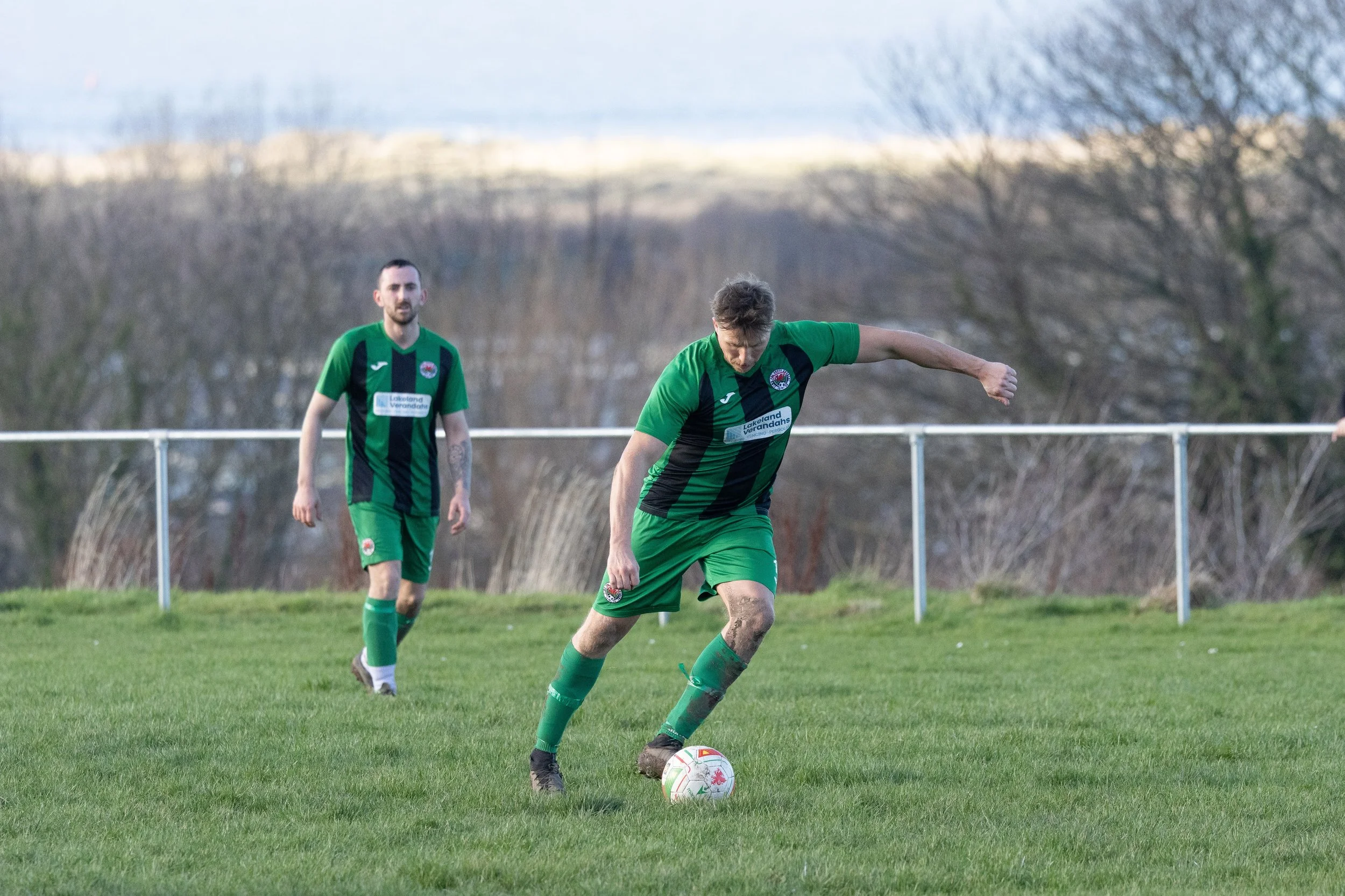 Two soccer players in green and black uniforms on a grass field, one preparing to kick a white soccer ball while the other stands in the background, with a fence and leafless trees in the distance.