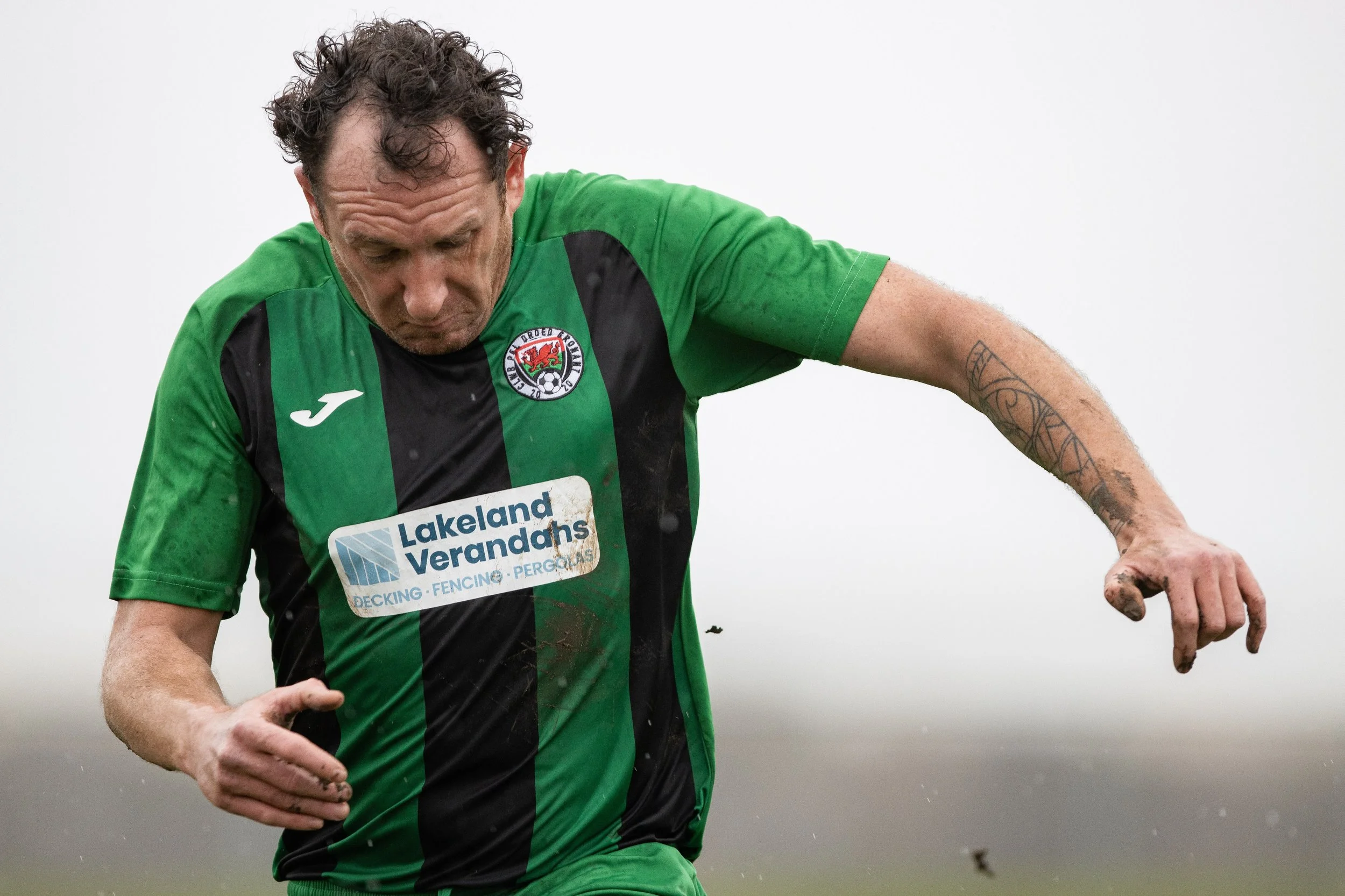 A soccer player in a green and black jersey with dirt on his arm and a tattoo on his forearm, running outdoors on a rainy day.